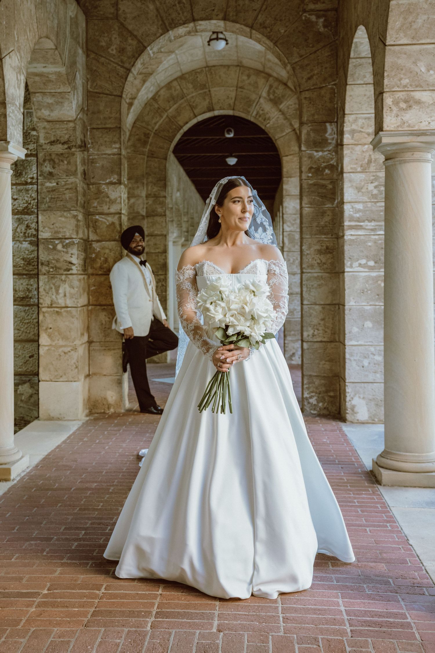 Bride in off-shoulder white ball gown with veil holds white bouquet under stone arches, groom in white suit behind.