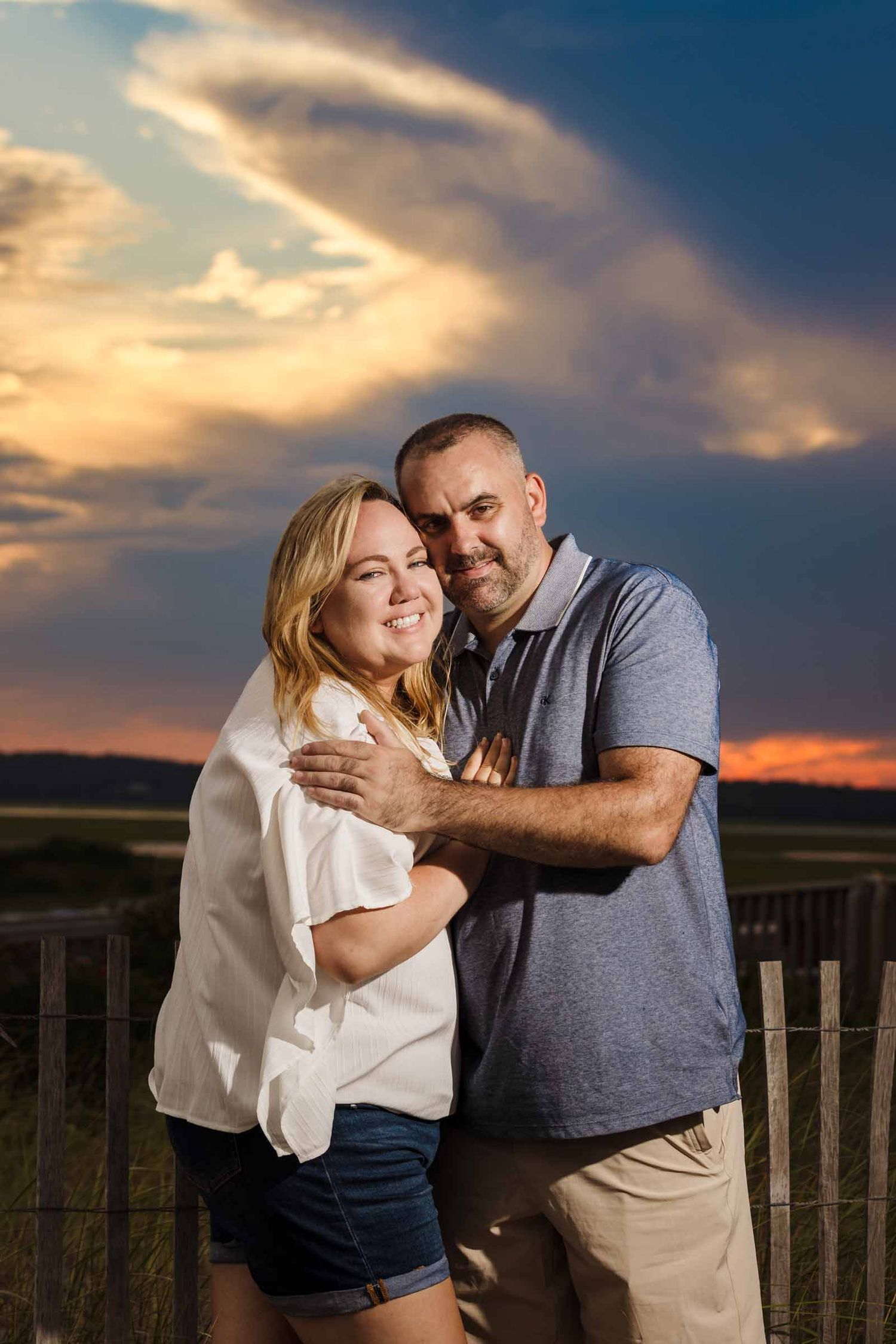 Engaged couple embracing by a wooden beach fence at Duxbury Beach, Massachusetts, under a dramatic orange and blue sunset.
