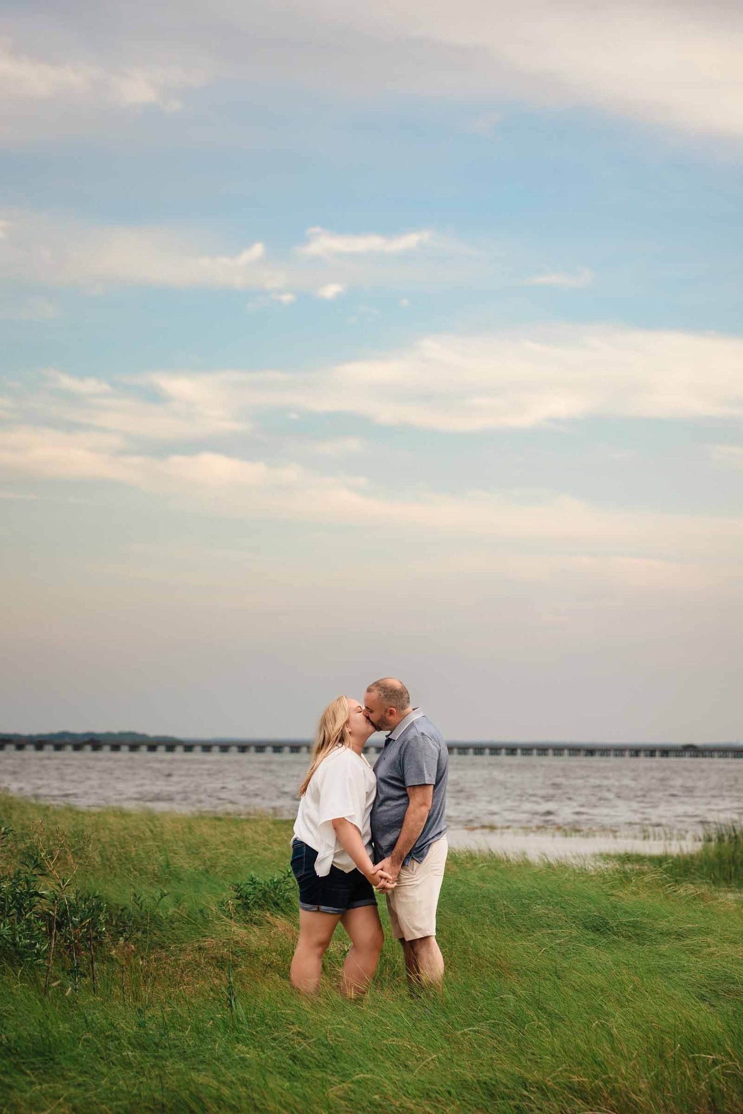 Engaged couple shares a kiss with Duxbury Beach in the background, the sky is a colorful sunset during their engagement session.