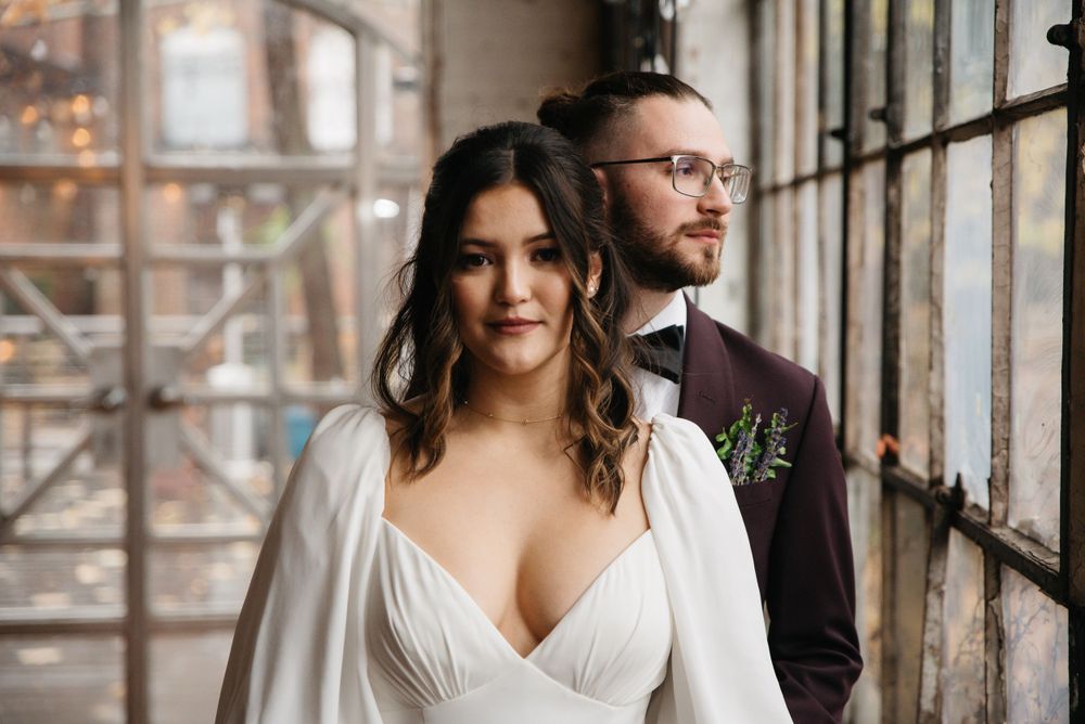 Elegant couple poses near industrial windows in wedding attire with beautiful natural lighting.