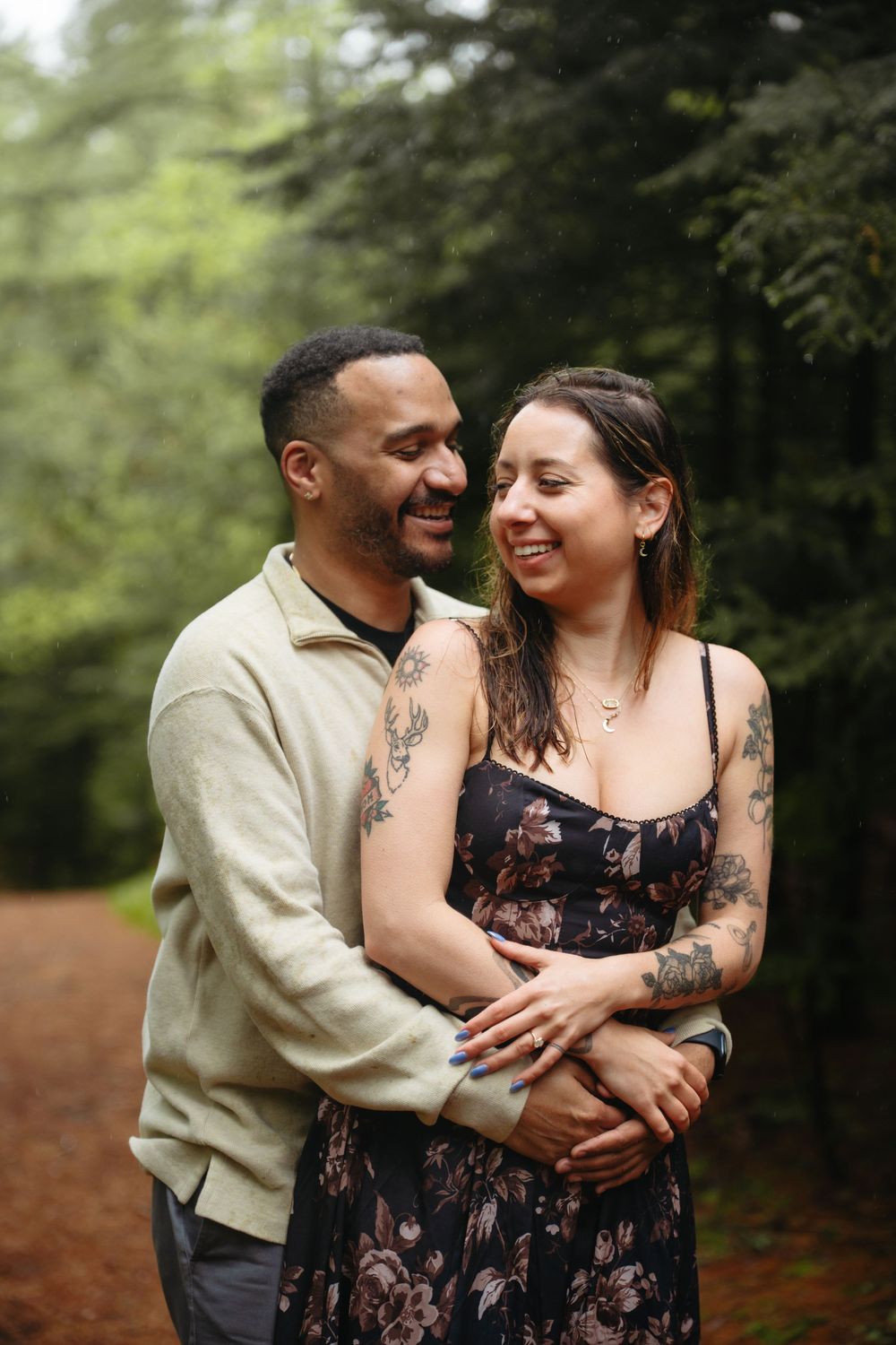 A smiling couple embraces on a forest path, the woman in a floral dress with tattoos, surrounded by lush green trees.
