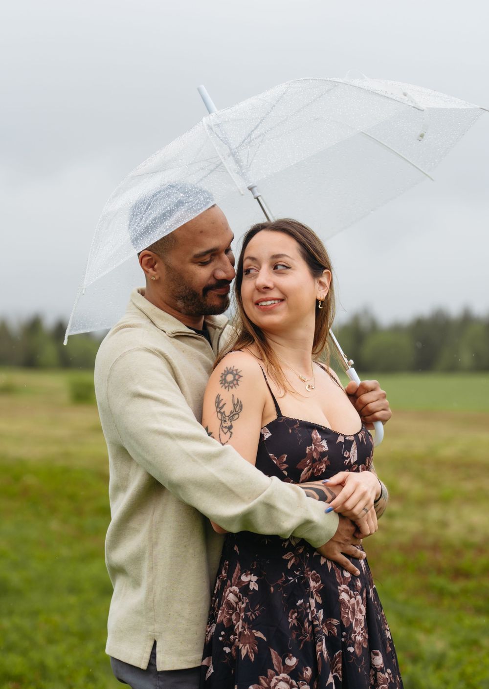 A couple shares a romantic embrace under a clear umbrella in a green field on a rainy day.