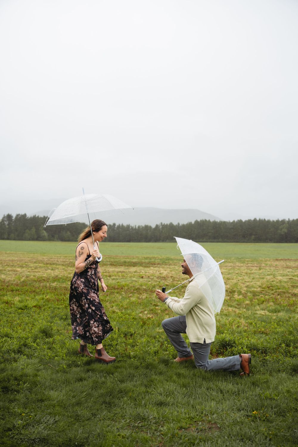 Man proposes on one knee in rainy green field, both holding clear umbrellas, woman in floral dress smiling joyfully.