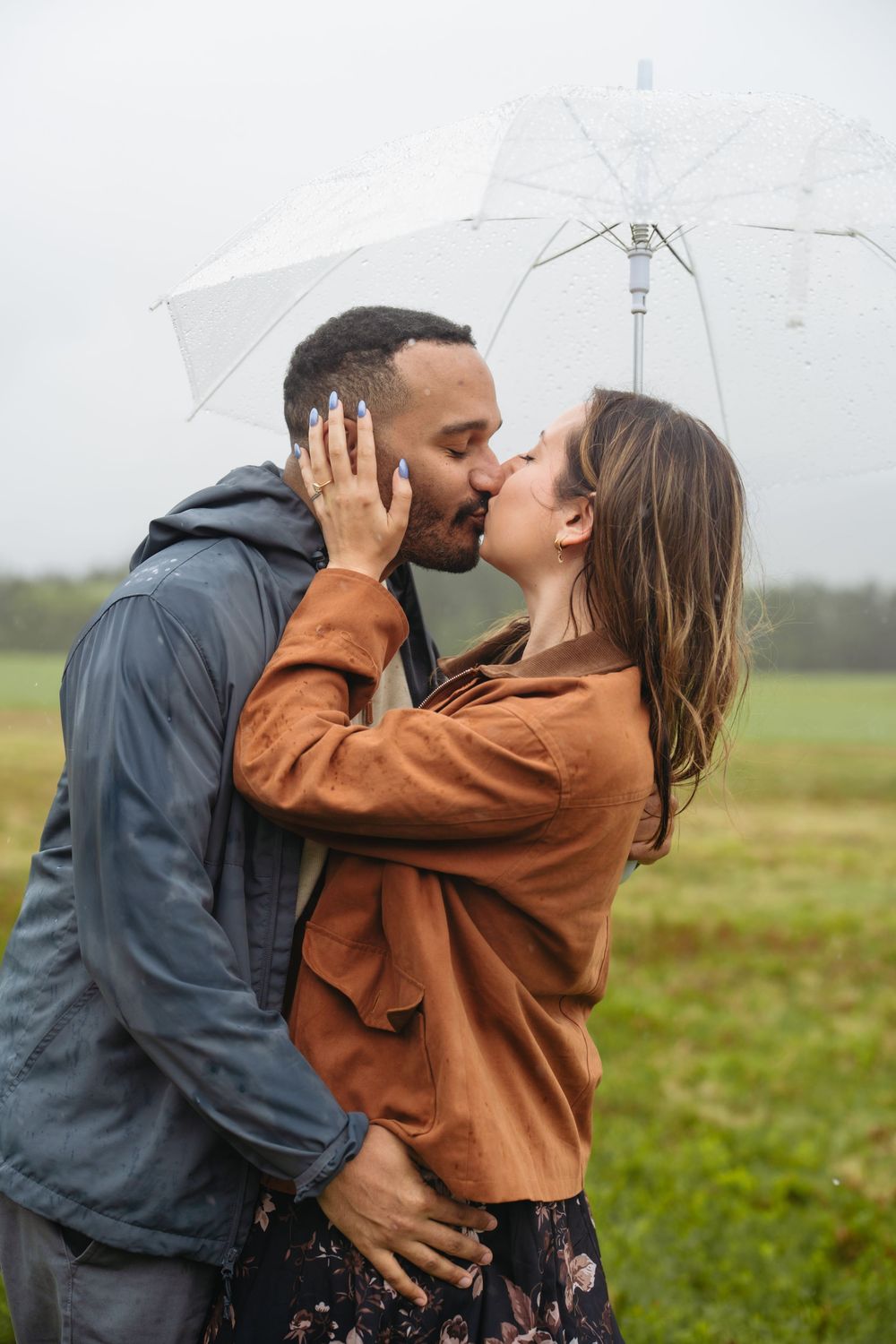 A romantic couple shares a kiss in the rain under a clear umbrella in a lush green field.