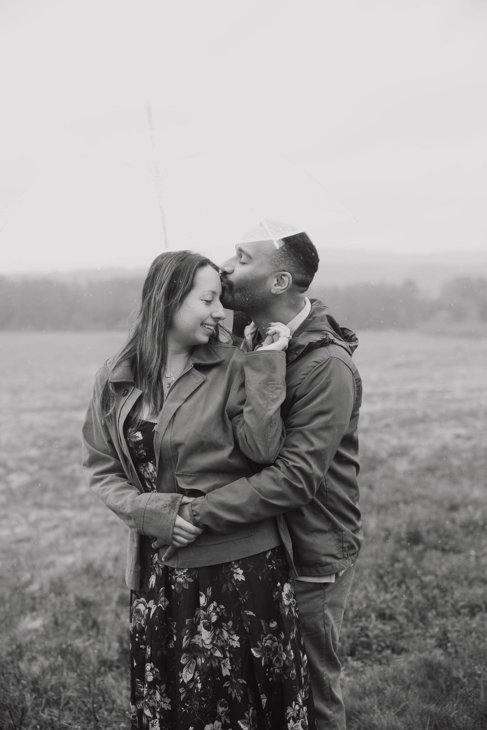 Black and white photo of a couple embracing in a field, man kissing woman on cheek, both wearing jackets outdoors.