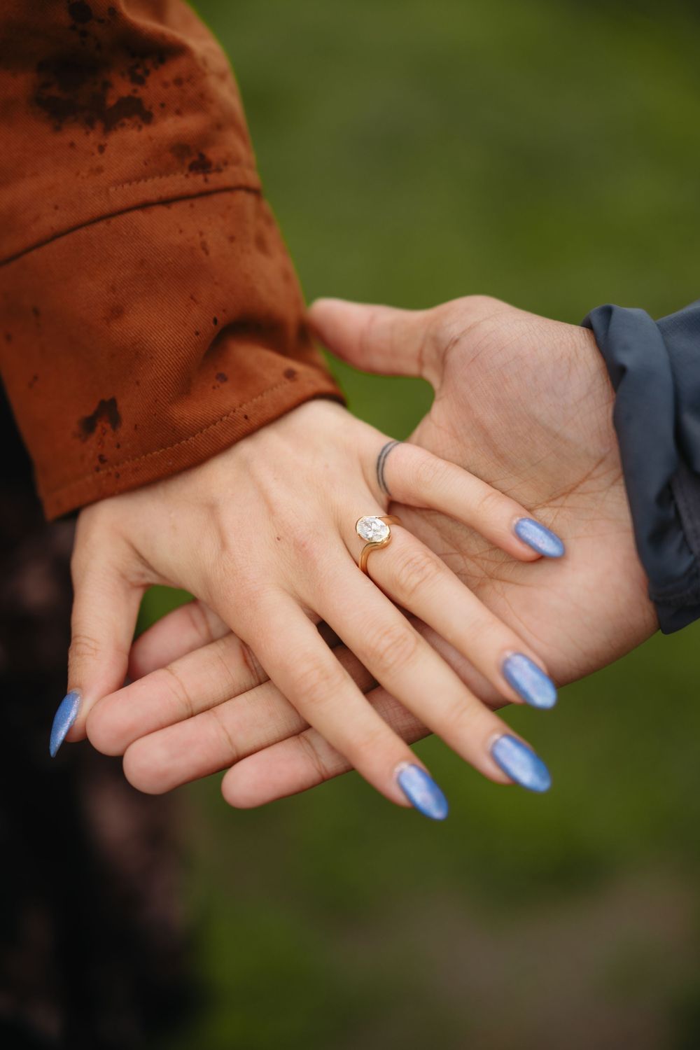 Close-up of two hands touching, woman wearing a diamond engagement ring with blue stiletto nails, man in gray sleeve.