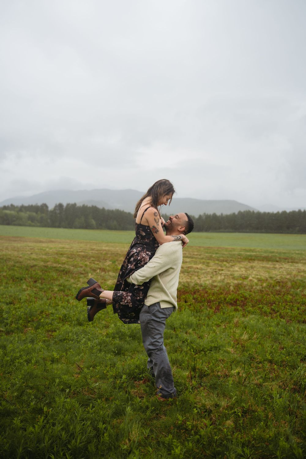 Man lifting woman in floral dress in green field with misty mountains, romantic outdoor couple photography.