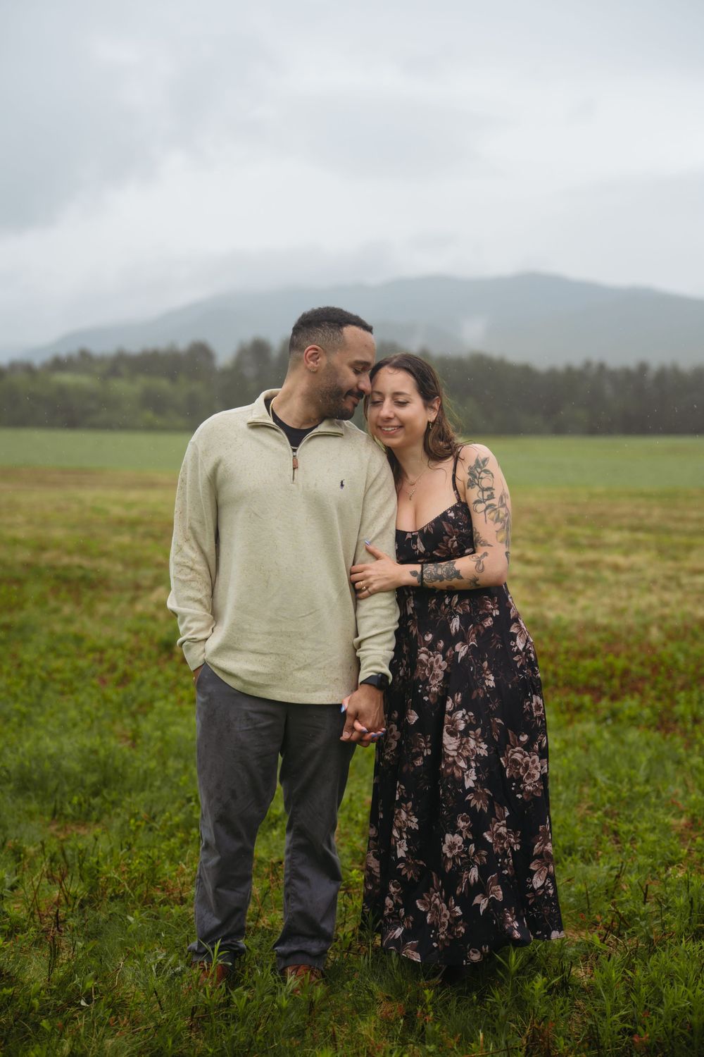 A couple holds hands and embraces in a lush green field with misty mountains, she wears a floral dress with tattoos.