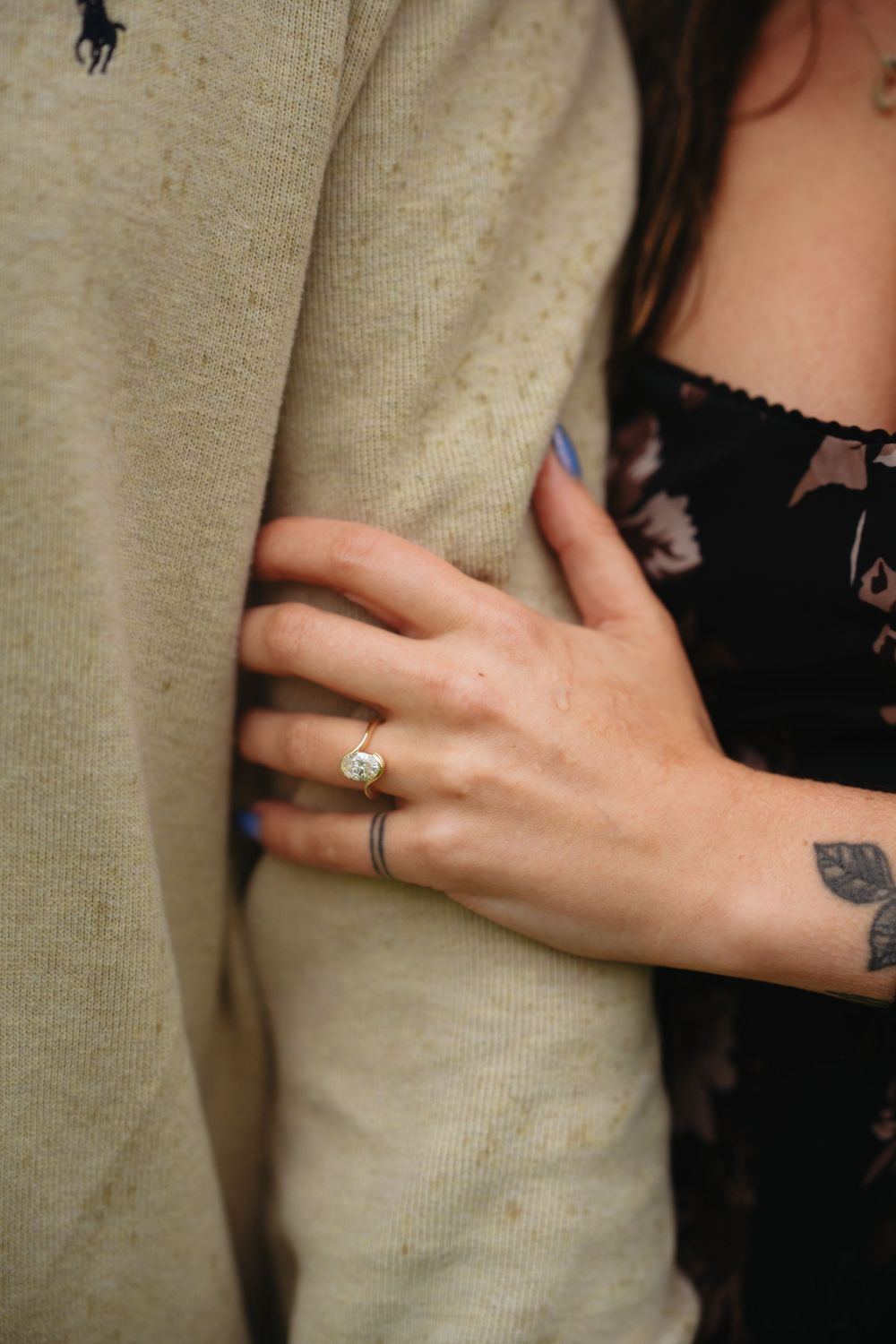Close-up of a woman's hand with an engagement ring and floral tattoo, resting on a man's arm in a beige sweater.