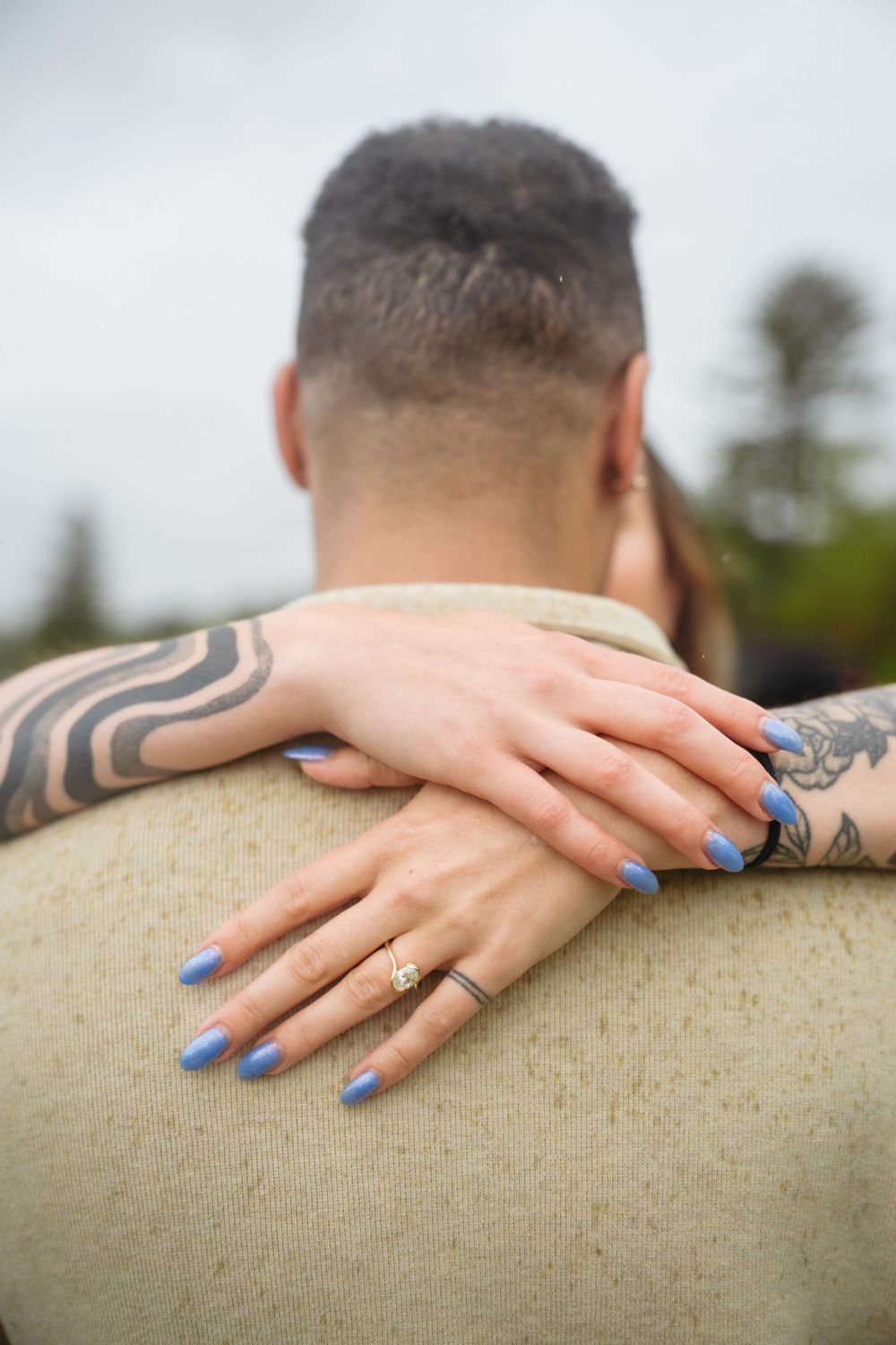 Close-up of a couple embracing, showing tattooed arms and blue manicured nails with an engagement ring.