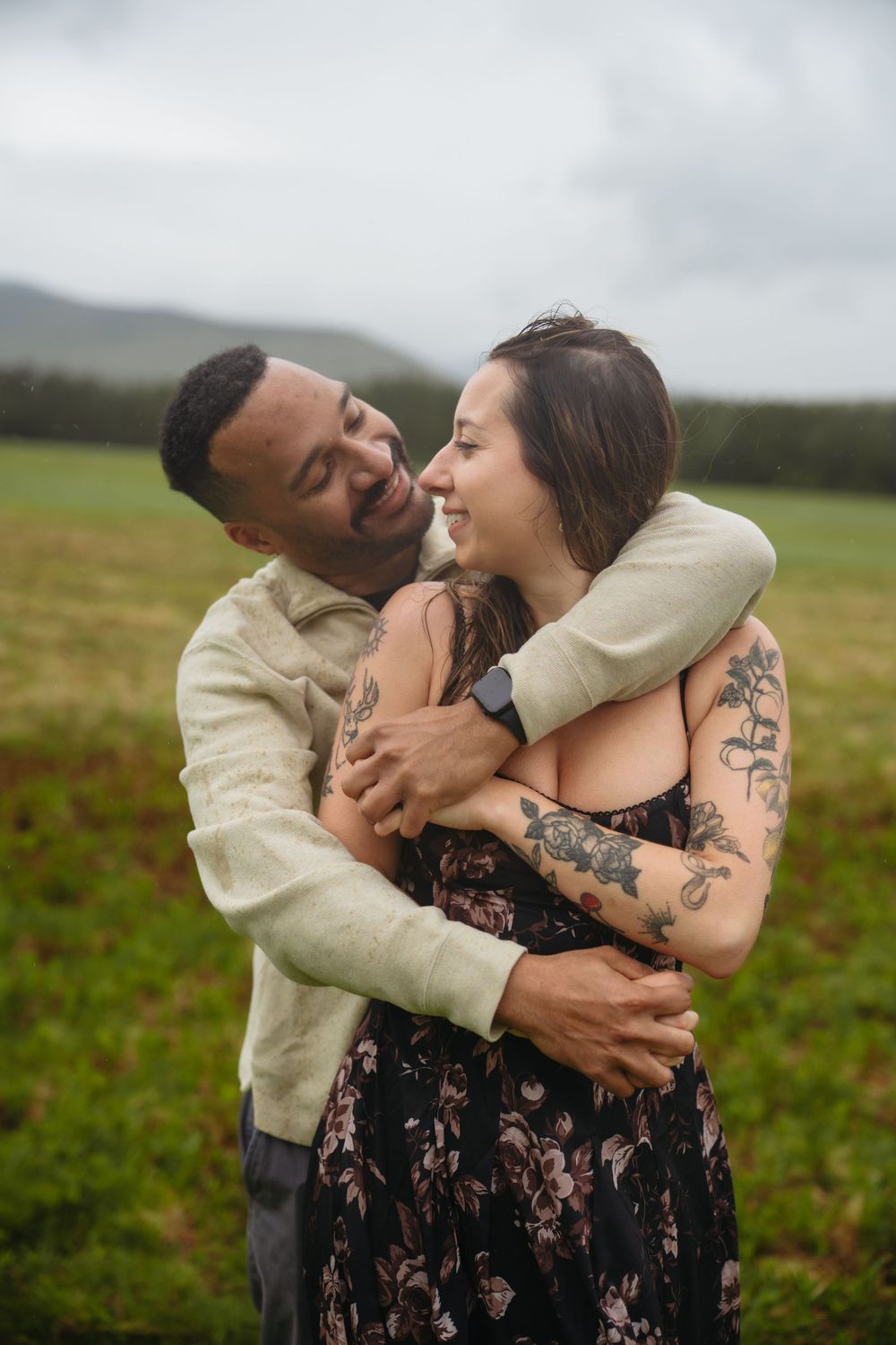 Smiling interracial couple embracing in a green field, woman wearing floral dress with tattoos.