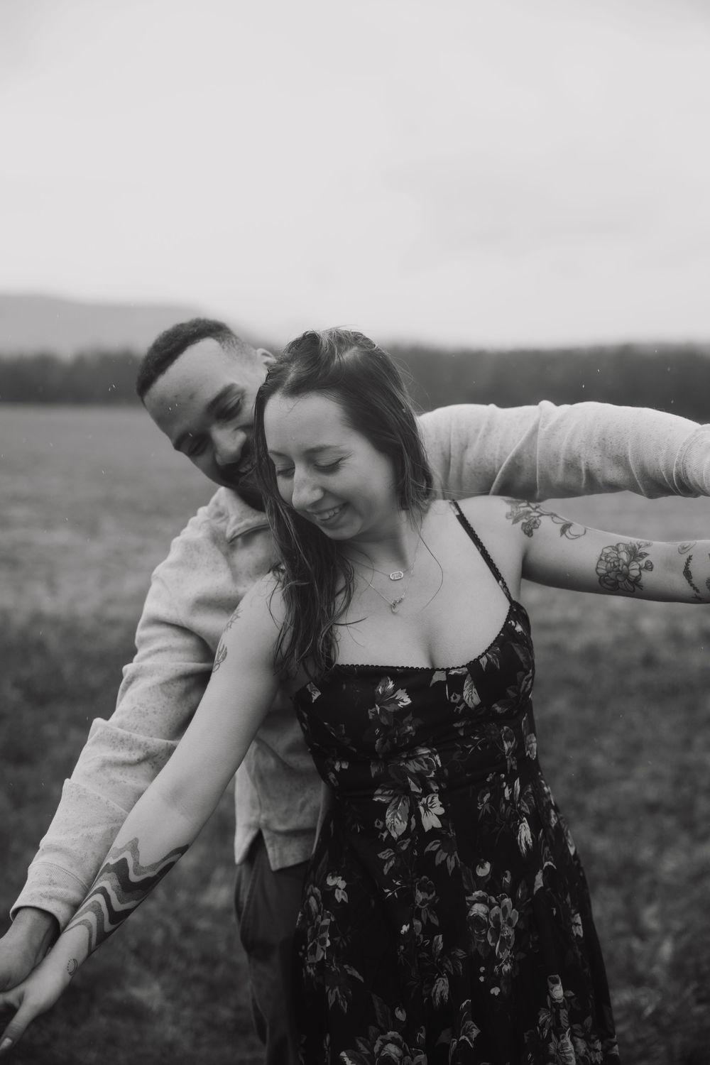 Black and white photo of a couple dancing joyfully in a field, woman in floral dress with tattoos.