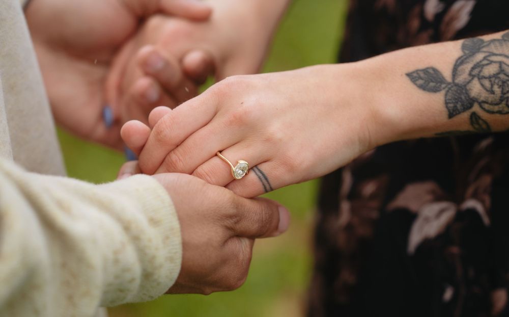 Close-up of two people holding hands, one wearing a pearl ring and floral tattoo on wrist, outdoors.