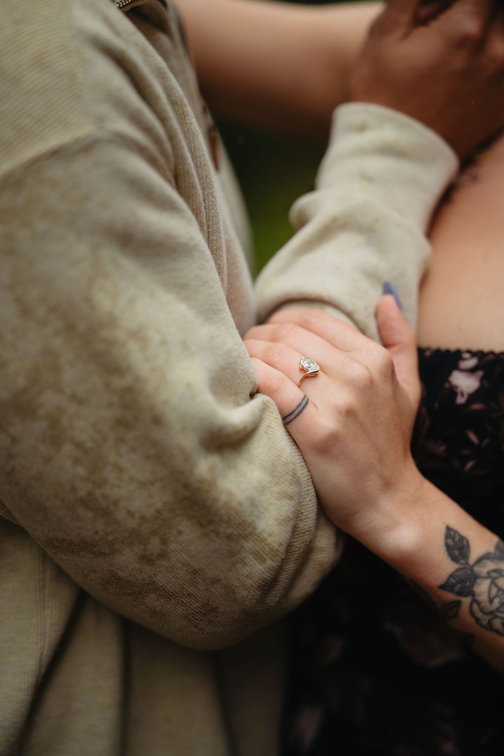 Close-up of a woman's hand with an engagement ring resting on a man's arm in a beige hoodie, with a floral tattoo visible.