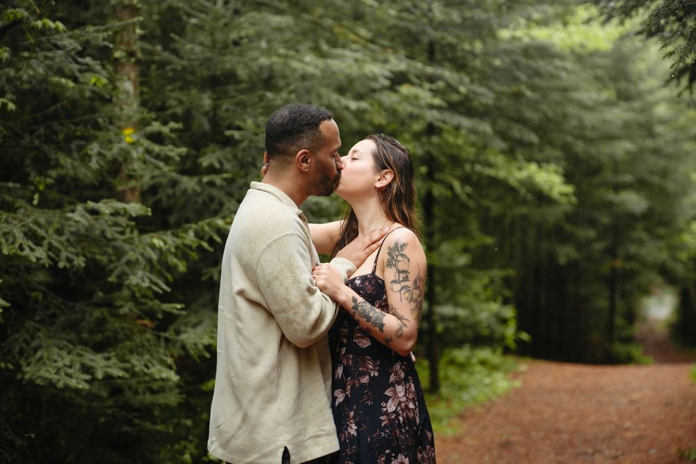 A couple shares a romantic moment in a lush forest, the woman in a floral dress with tattoos leaning in to kiss her partner.