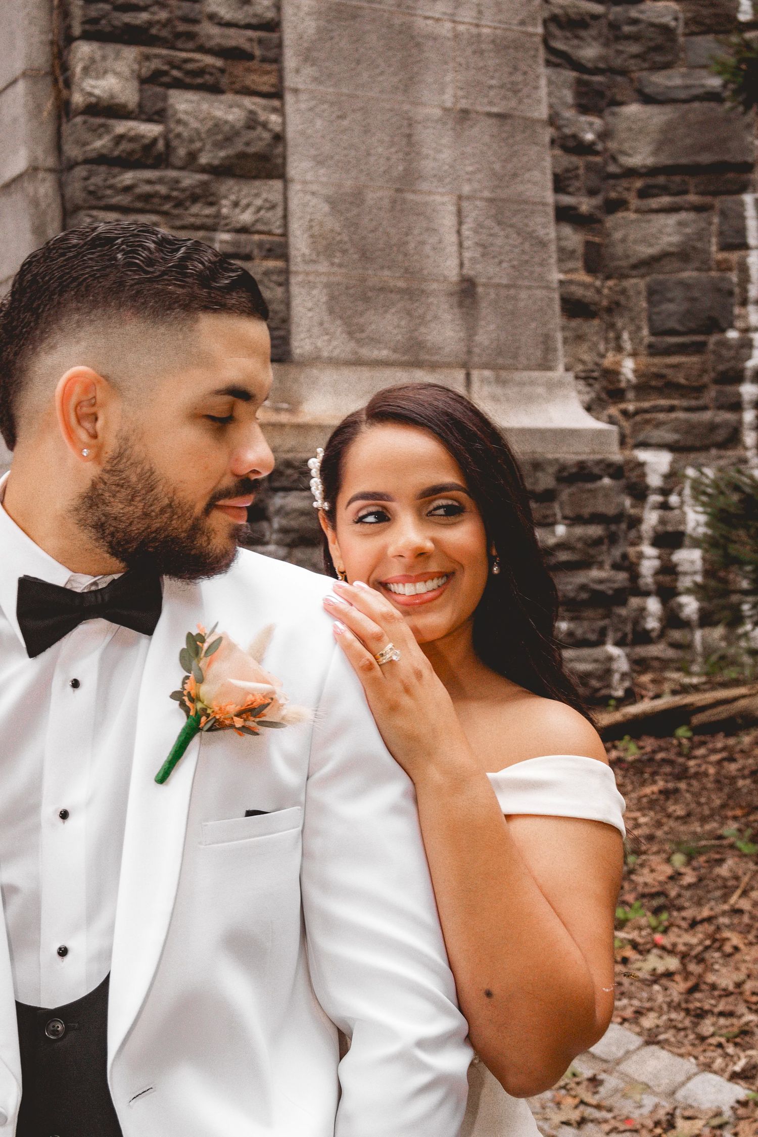A bride and groom pose together in wedding attire, she smiles at camera while he gazes at her near a stone building.