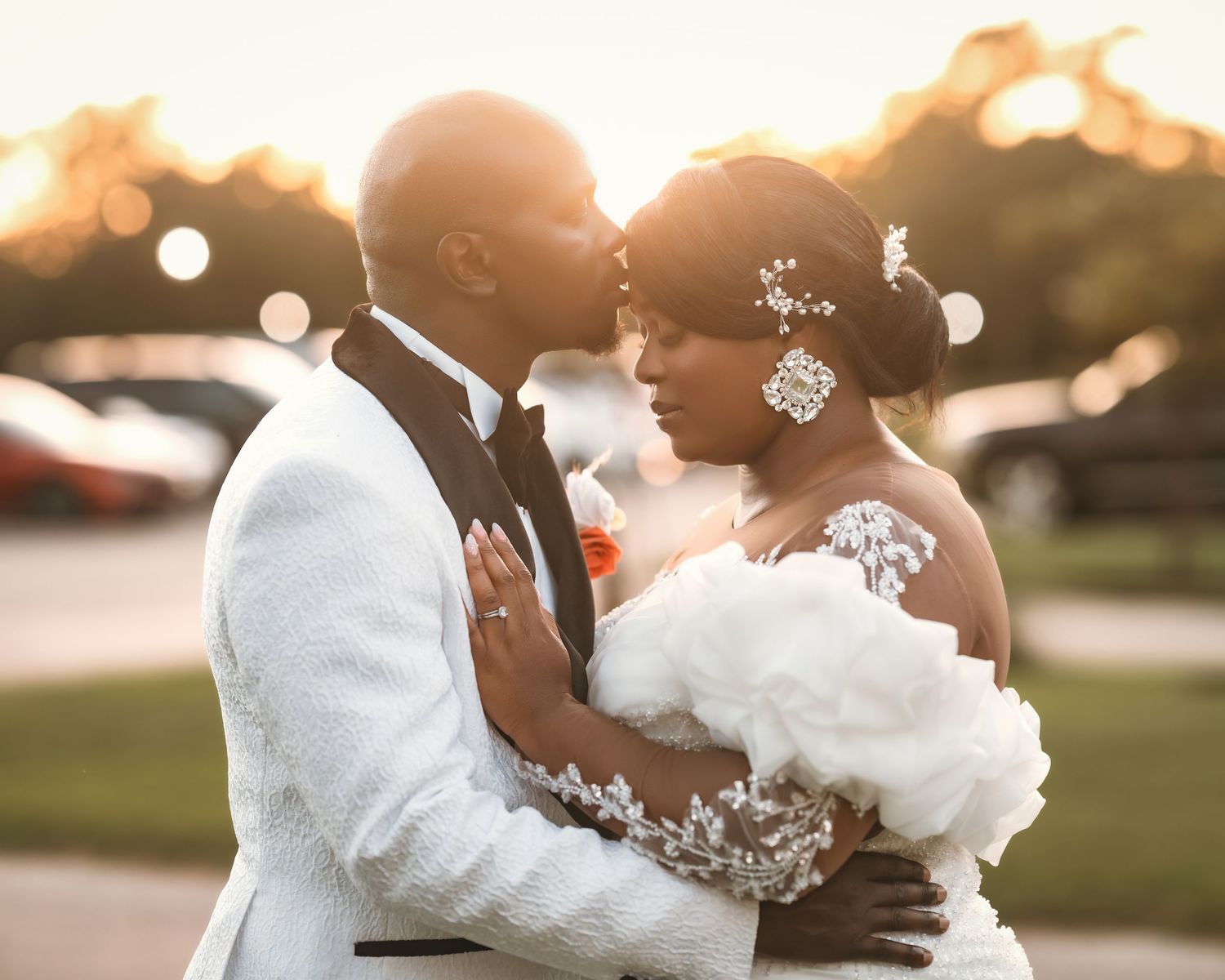 A Black couple shares a romantic embrace at golden hour, bride in white gown with floral hairpiece, groom in white suit.