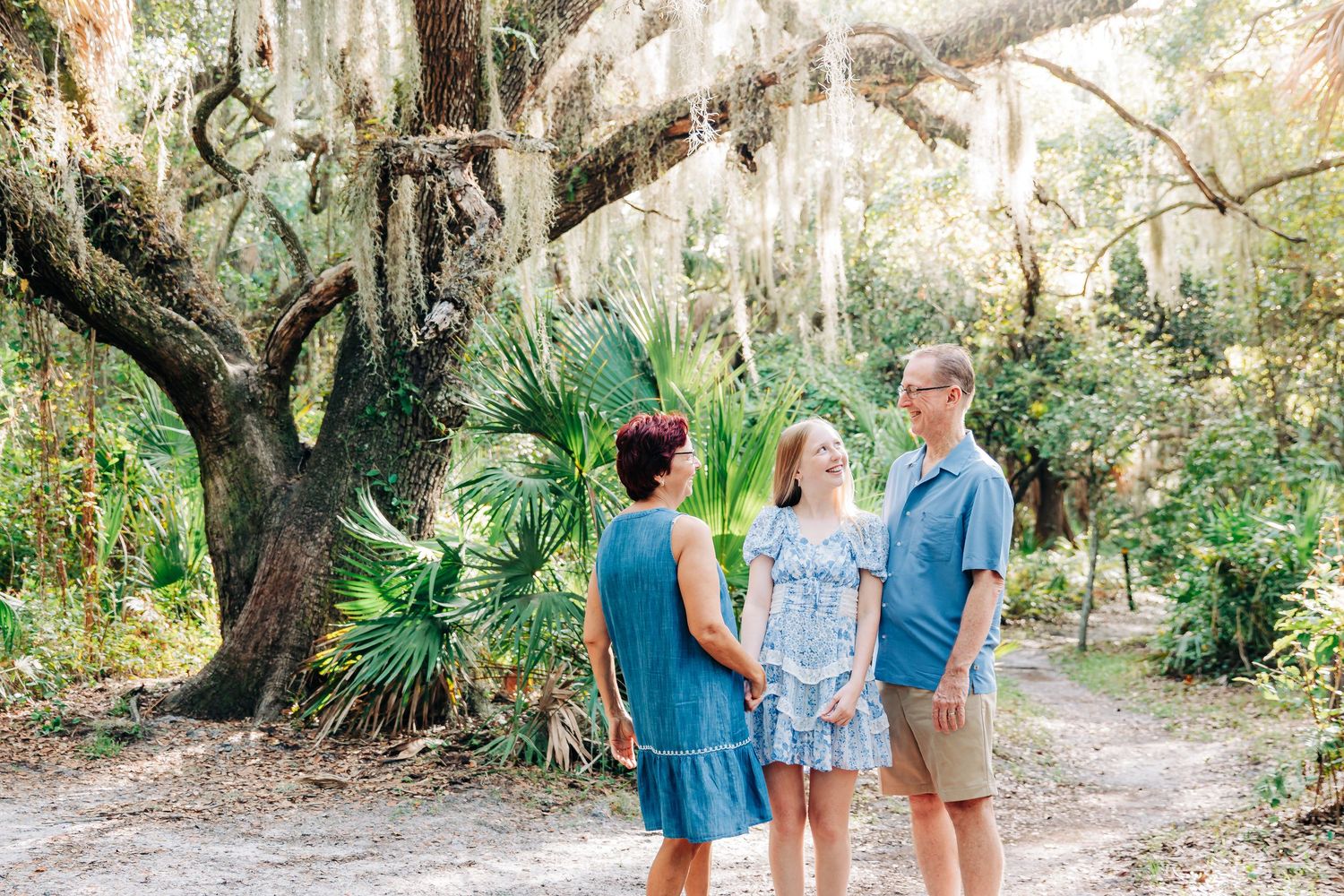 Family of three poses on a sandy path surrounded by lush greenery and Spanish moss-draped oak trees at Delray Oaks Natural Area.