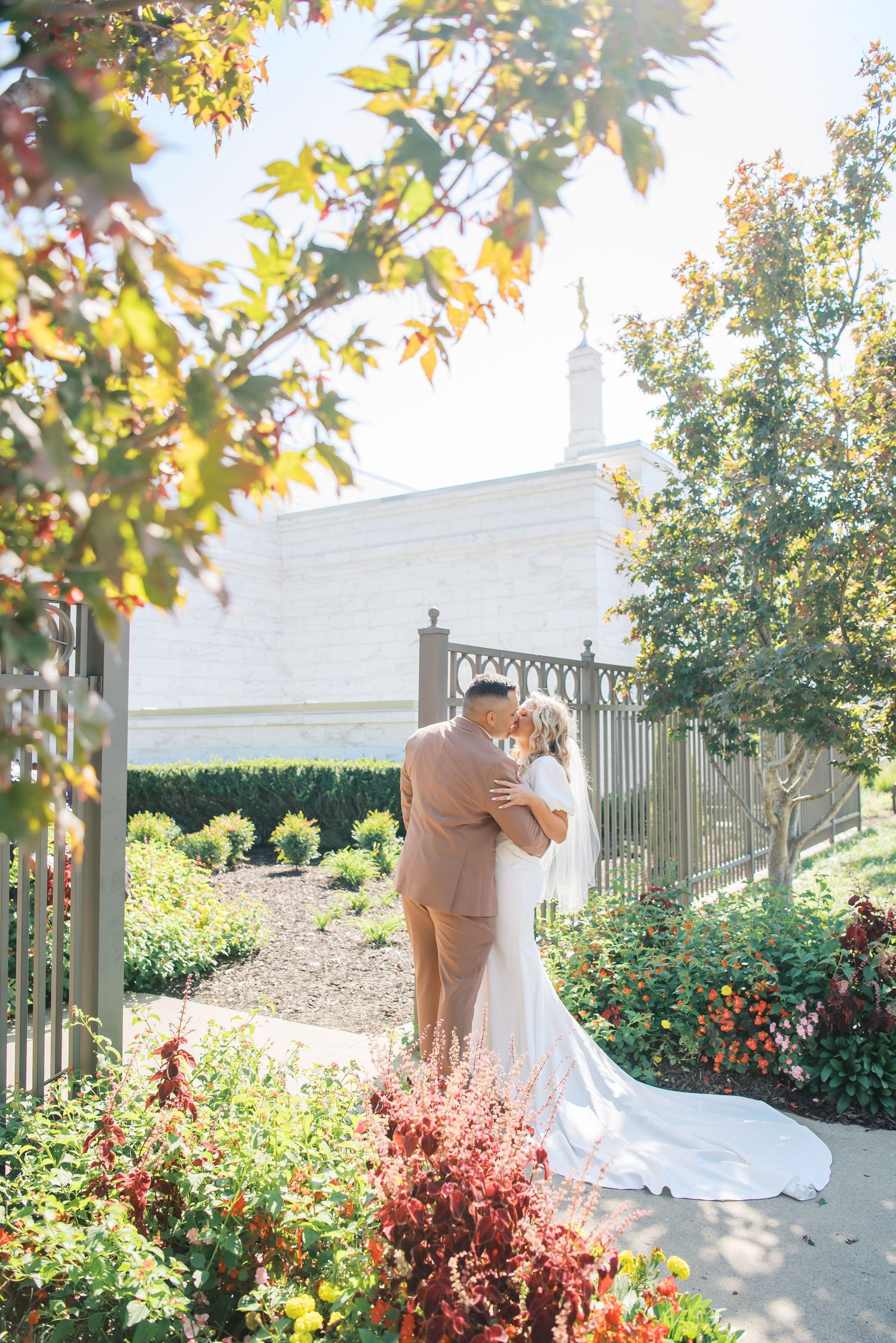 Bride and groom share a romantic kiss in a lush garden near a white building with an iron gate on their wedding day.