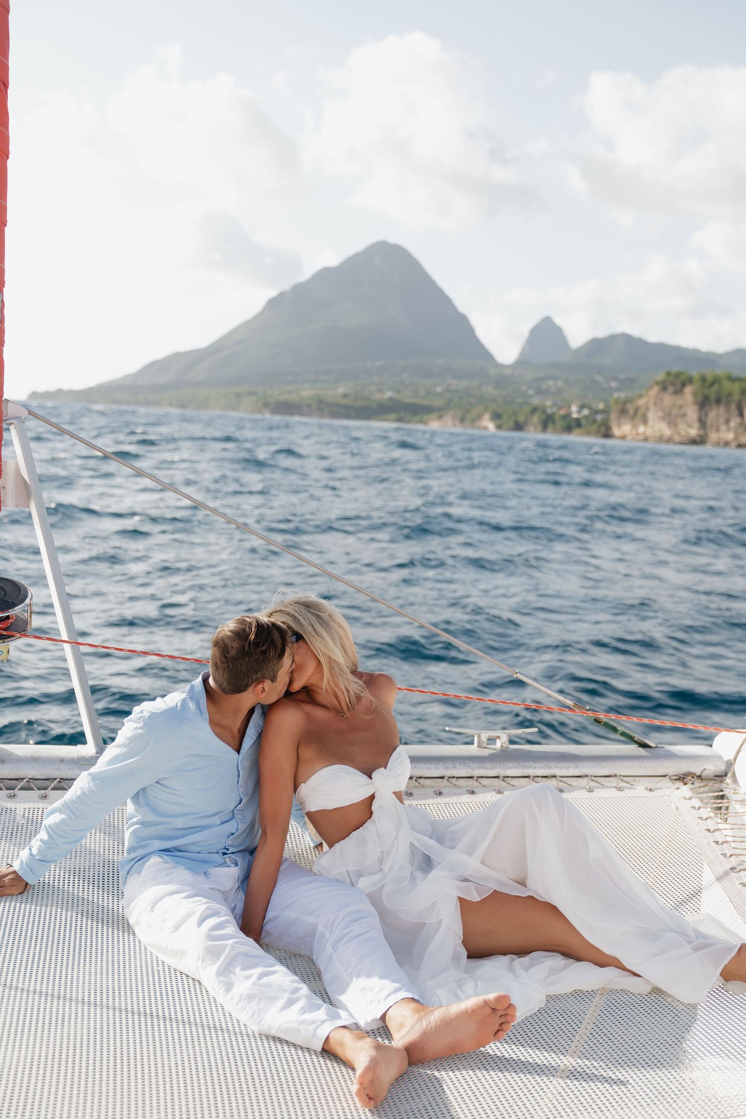 Romantic couple kissing on a luxury catamaran sailboat deck with stunning mountain island views in the background.