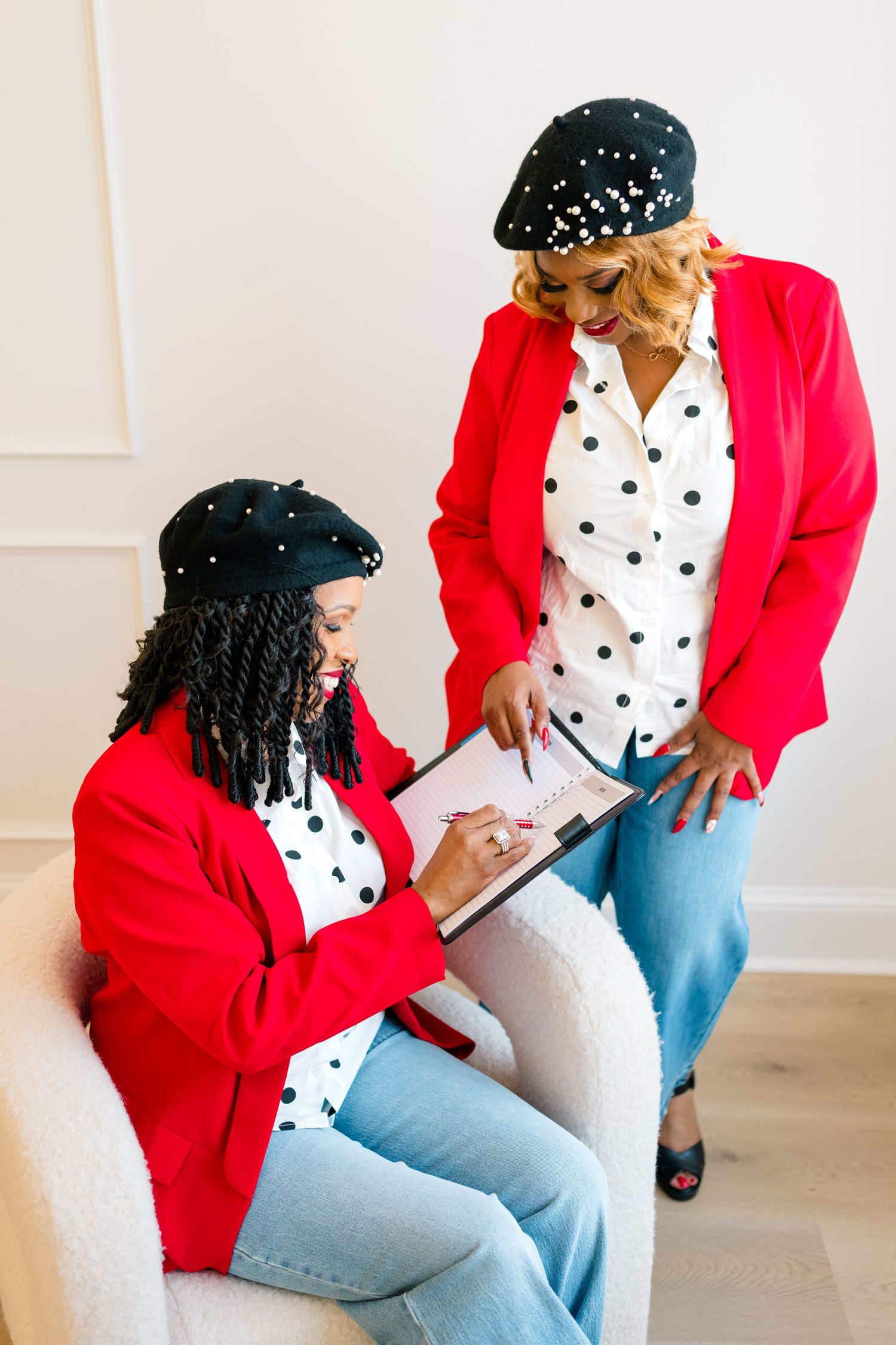 Two women in matching red blazers, polka dot blouses, jeans, and black berets review notes together in a bright studio.