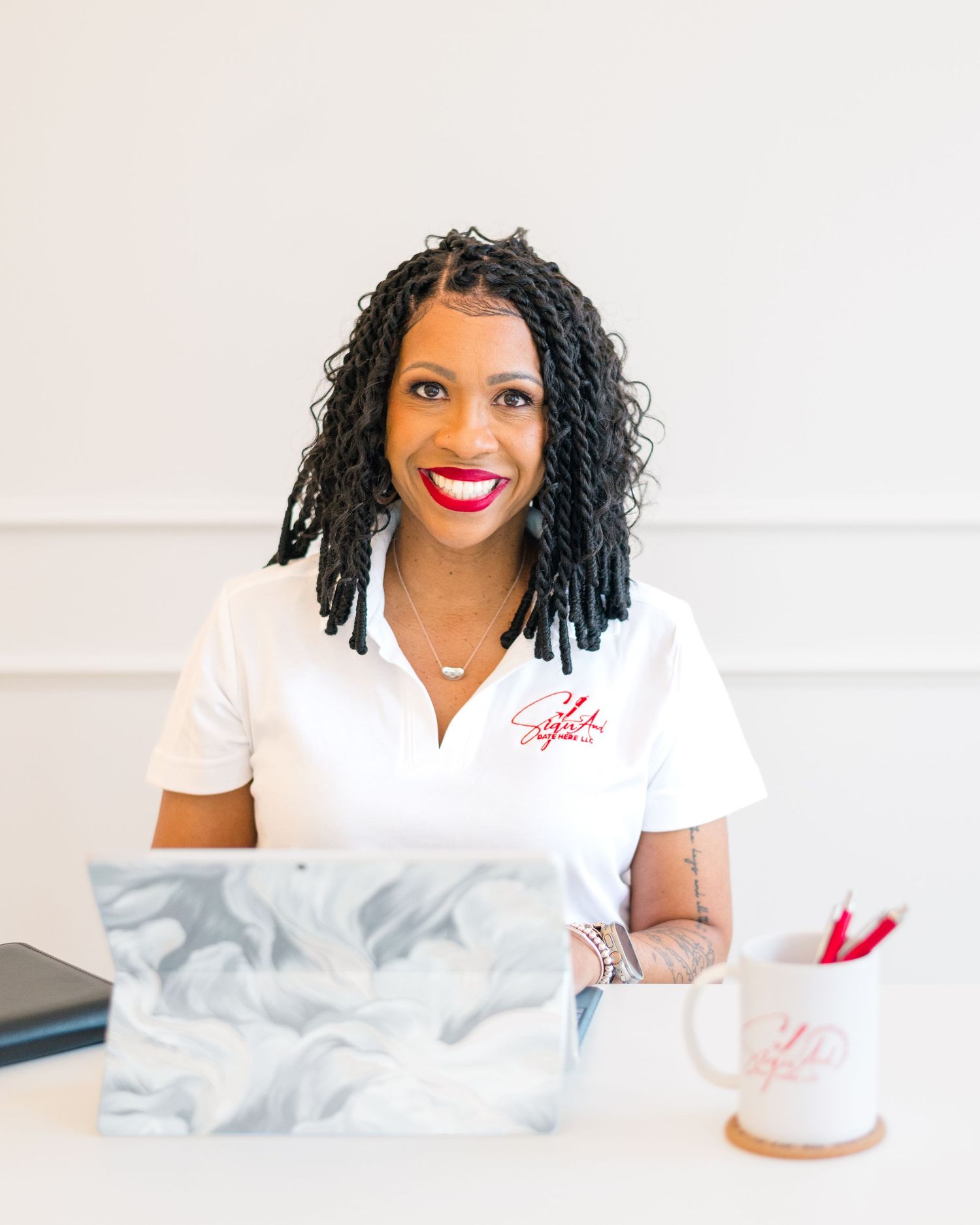 Smiling woman with locs and red lipstick in white polo sits at desk with marble laptop and coffee mug.