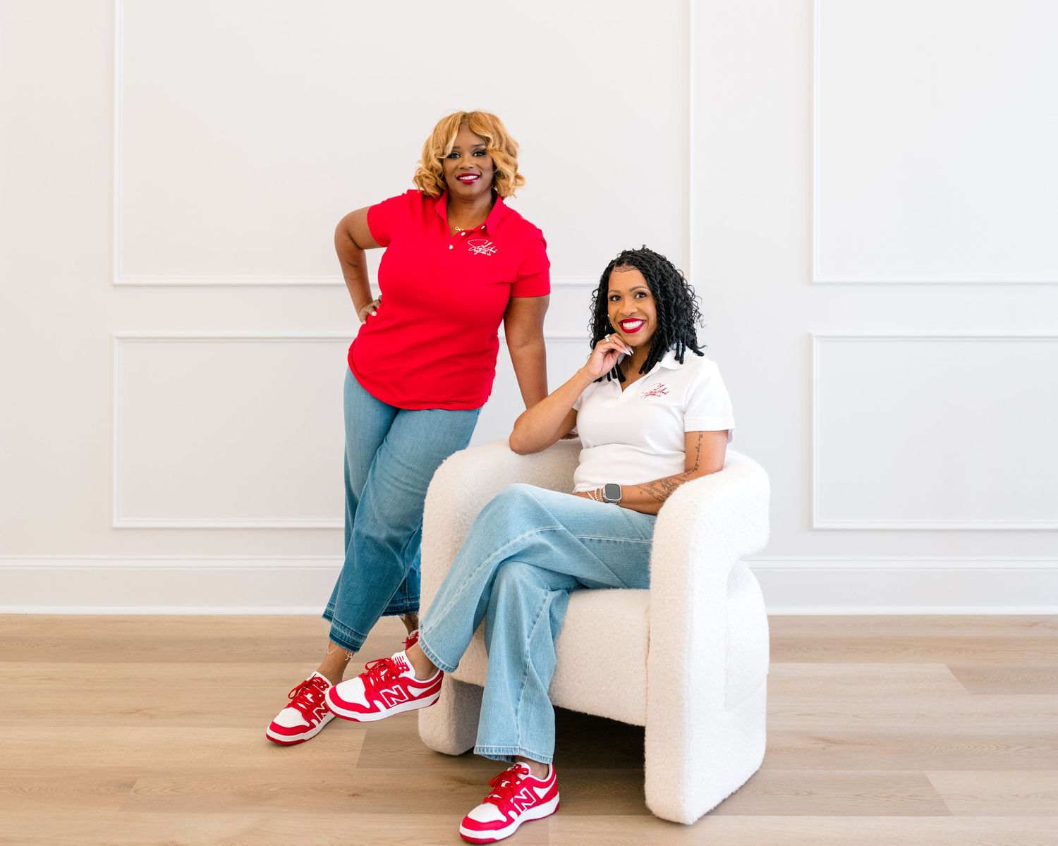 Two women in red and white outfits pose in a bright studio, one seated on a white chair, both wearing red sneakers.