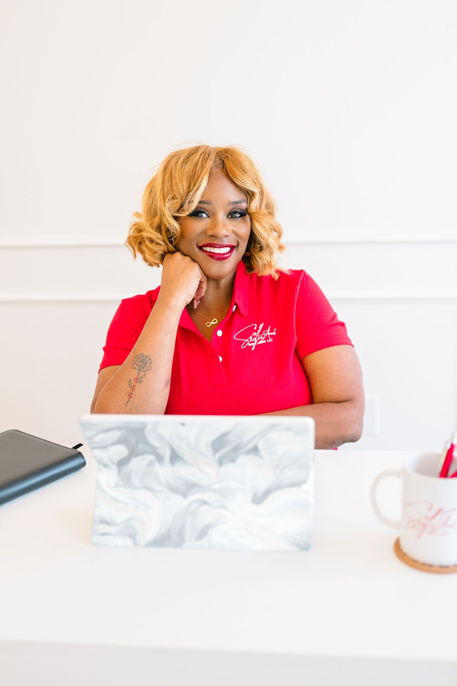 Smiling woman in red polo shirt sitting at white desk with marble laptop, bright professional office setting.