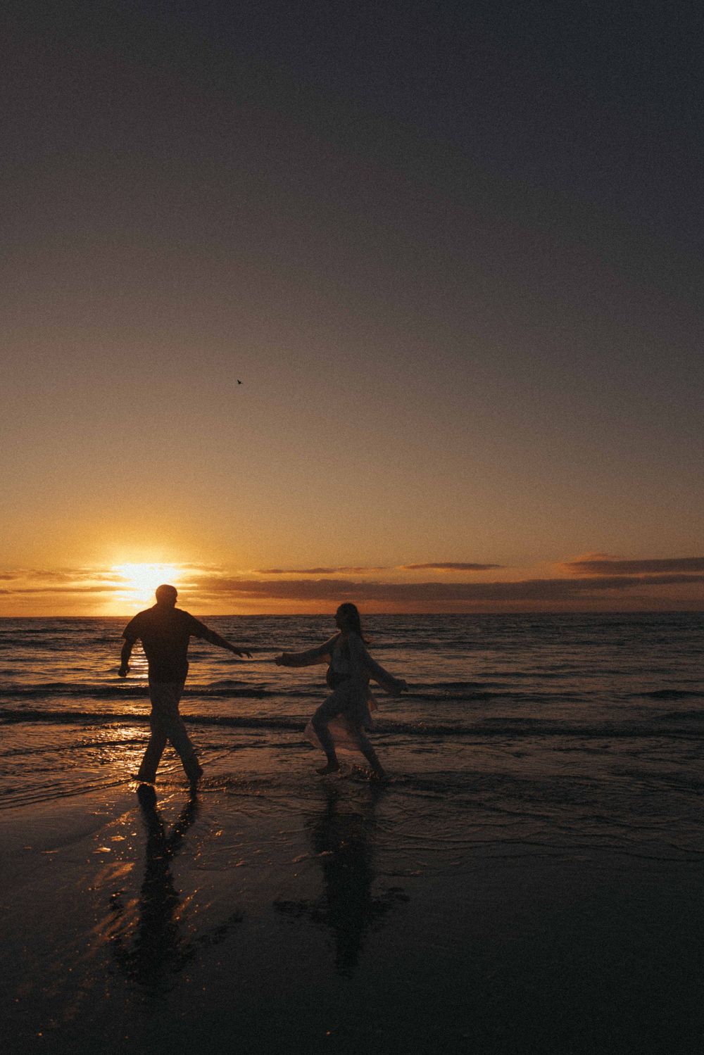 Silhouette of a couple running on a beach at sunset with golden reflections on wet sand.