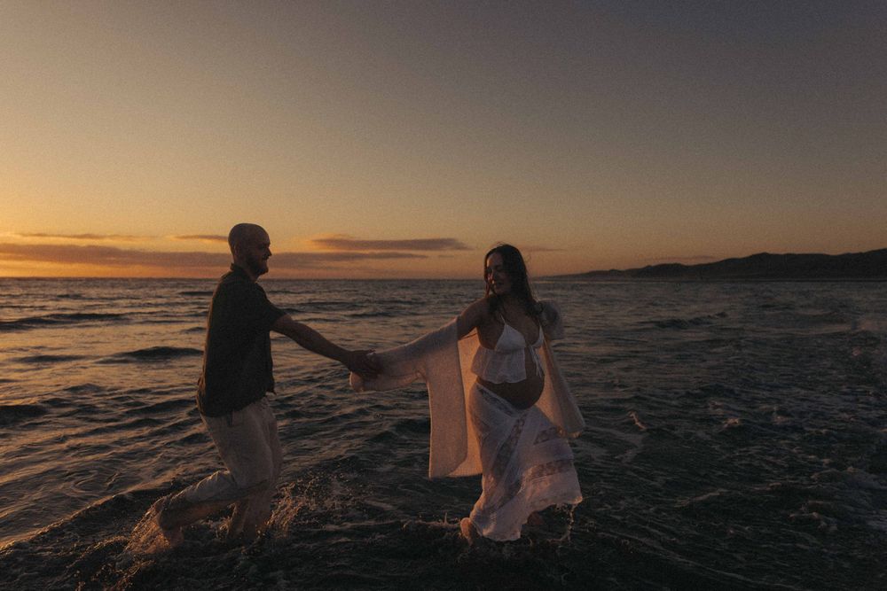 Couple holding hands wading in ocean waves at golden sunset, woman in white dress, romantic beach engagement photo.