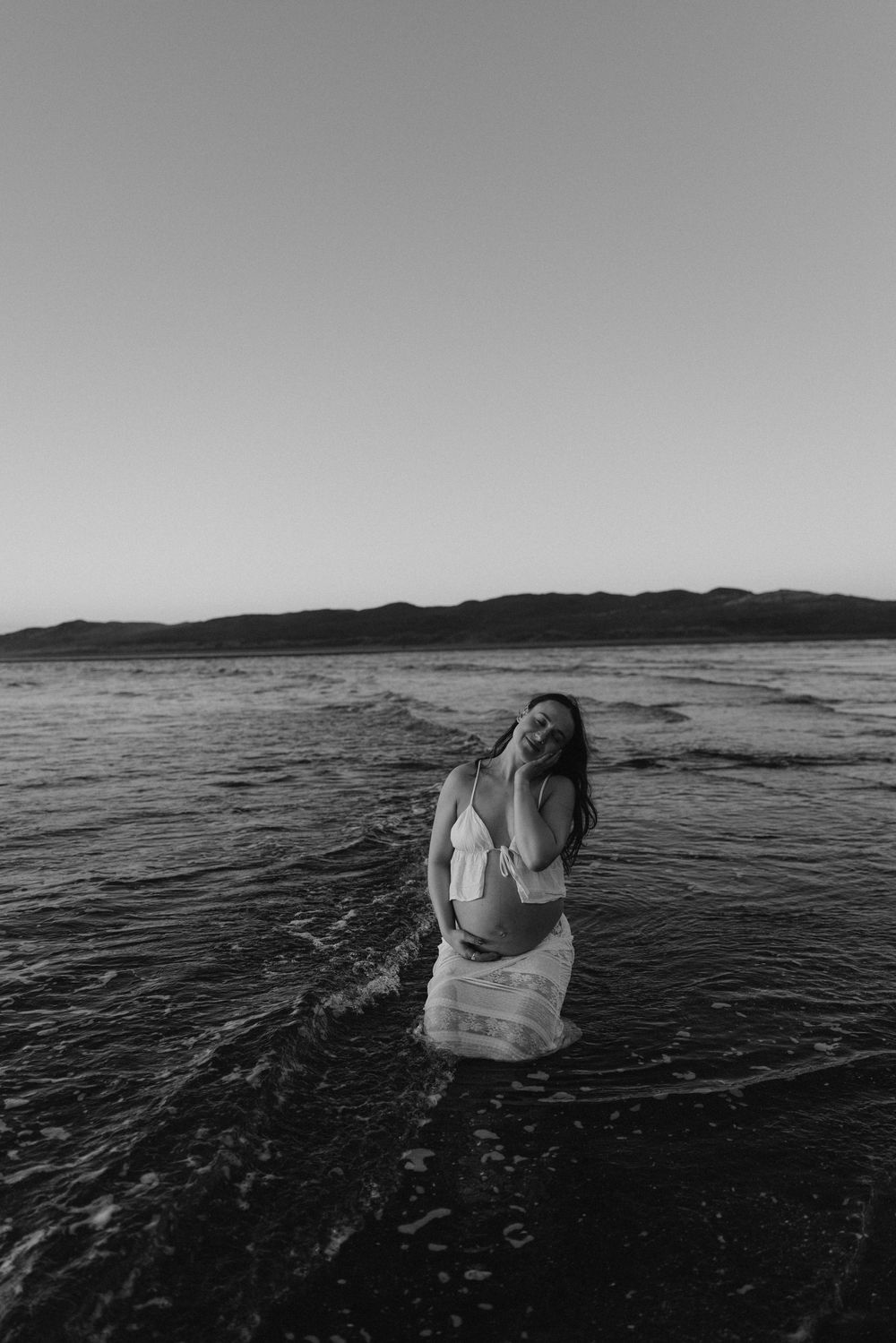 Black and white photo of a woman standing in ocean waves wearing a white dress, with mountains in the background.