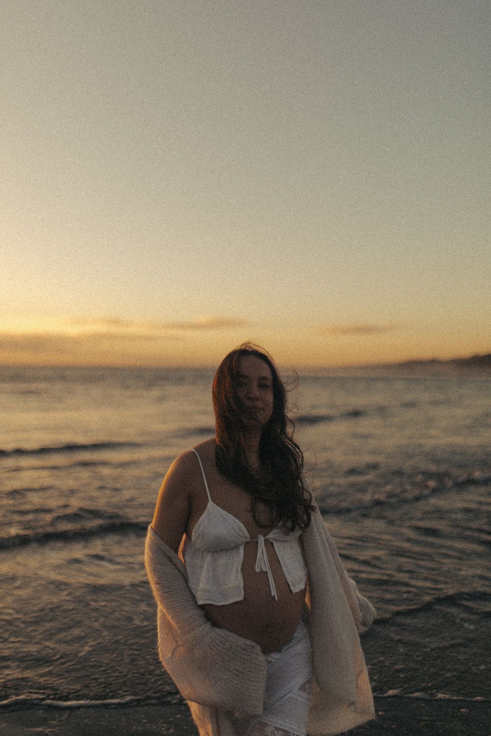 Pregnant woman in white bikini and cardigan standing on beach at golden hour sunset.