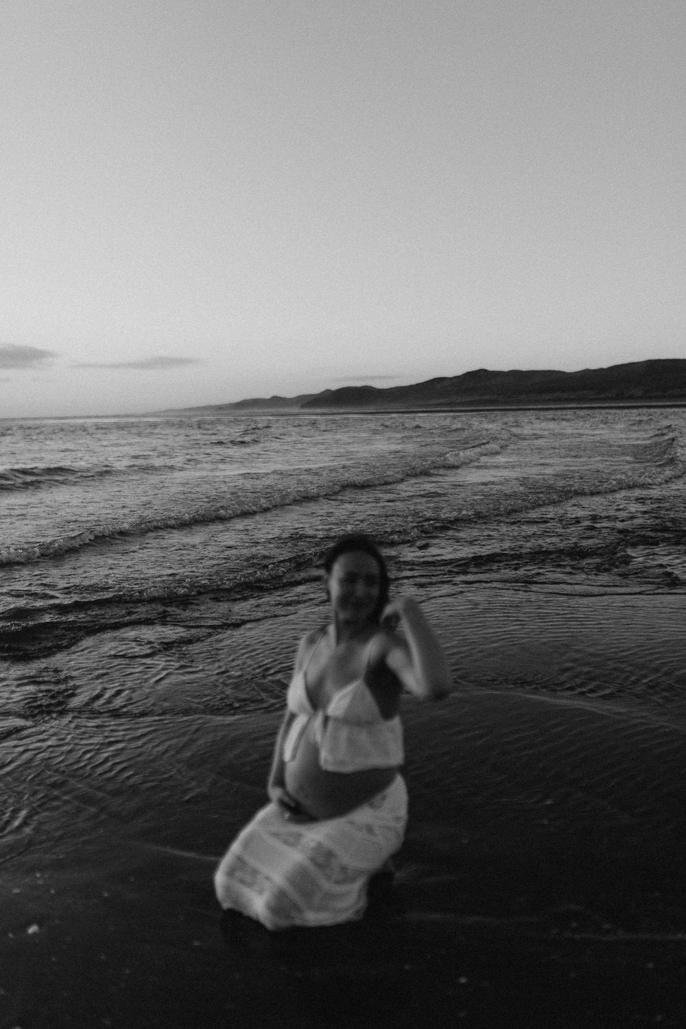 Black and white photo of a pregnant woman standing in ocean waves at dusk on a serene beach.
