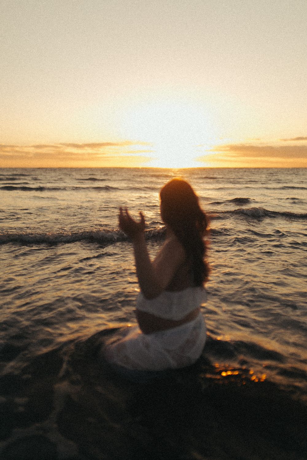 Woman in white dress sitting in ocean waves at golden sunset, viewed from behind with sun on horizon.