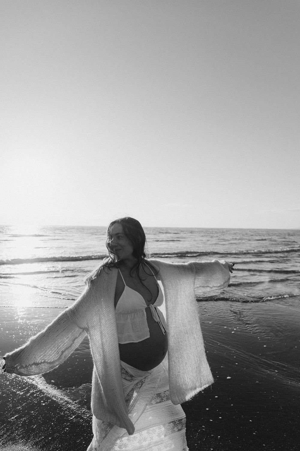 Black and white photo of a pregnant woman in a bikini and flowing cover-up posing on a sunny beach shoreline.