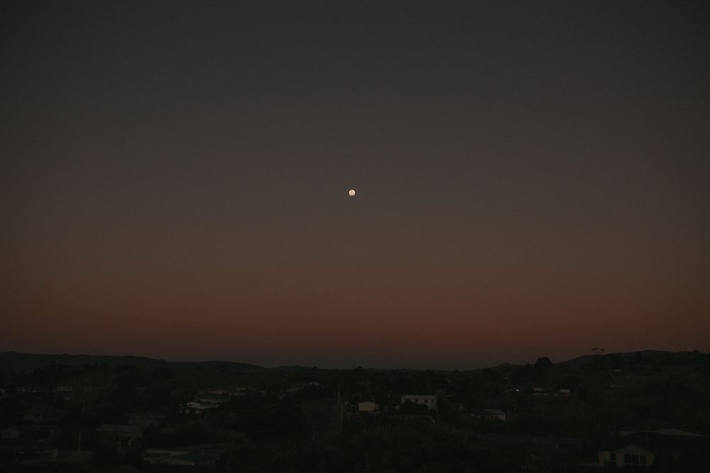 Twilight sky with a crescent moon glowing above a dark cityscape silhouette at dusk.
