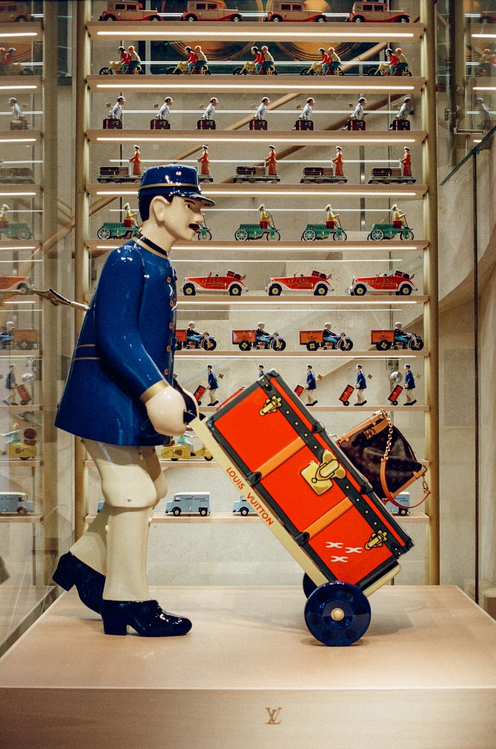 Louis Vuitton bellhop figurine with orange LV trunk on dolly, displayed in store with vintage toy figurines.