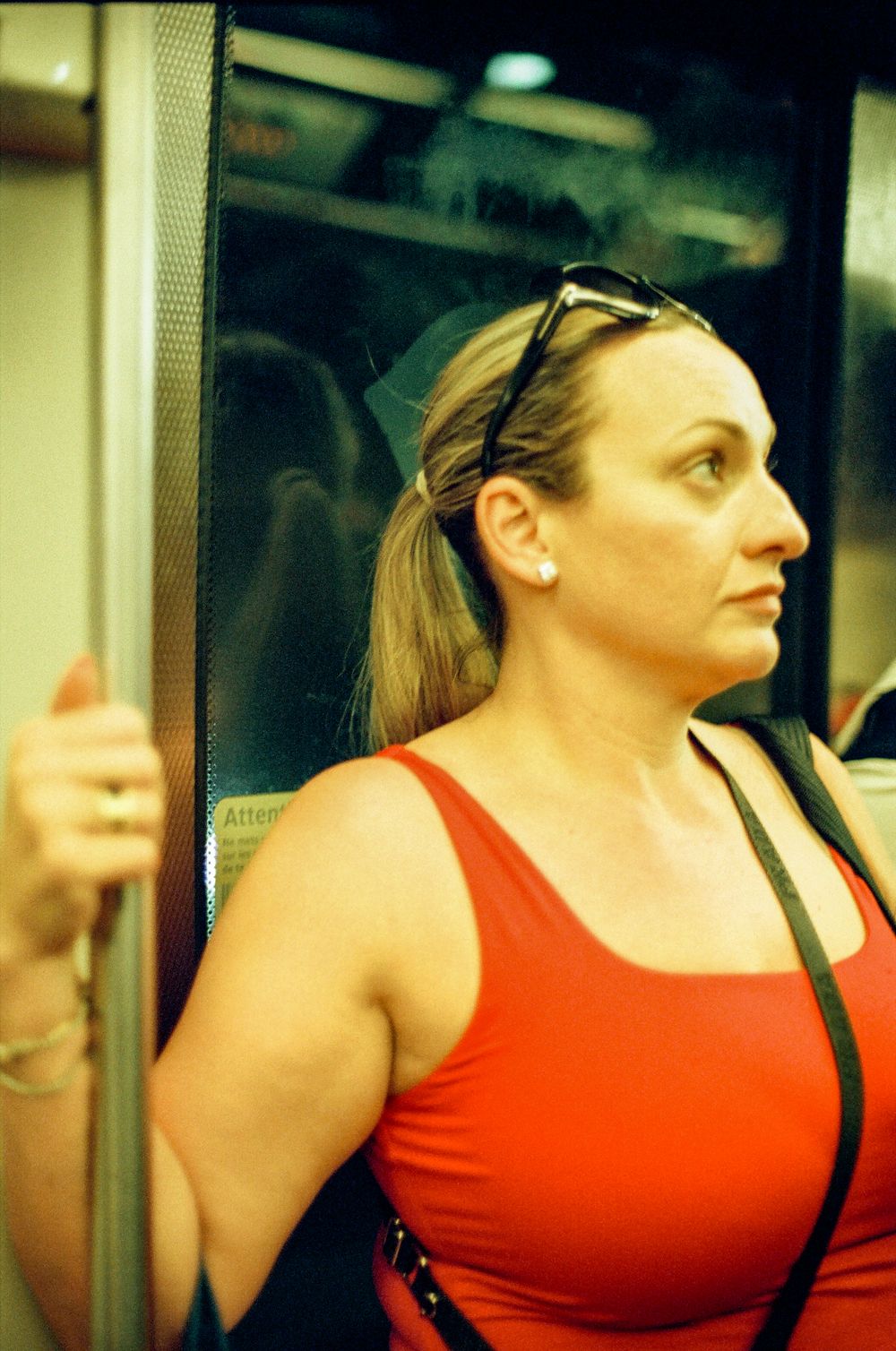 Woman in red tank top holding subway pole, sunglasses on head, wearing earrings and bag strap visible.