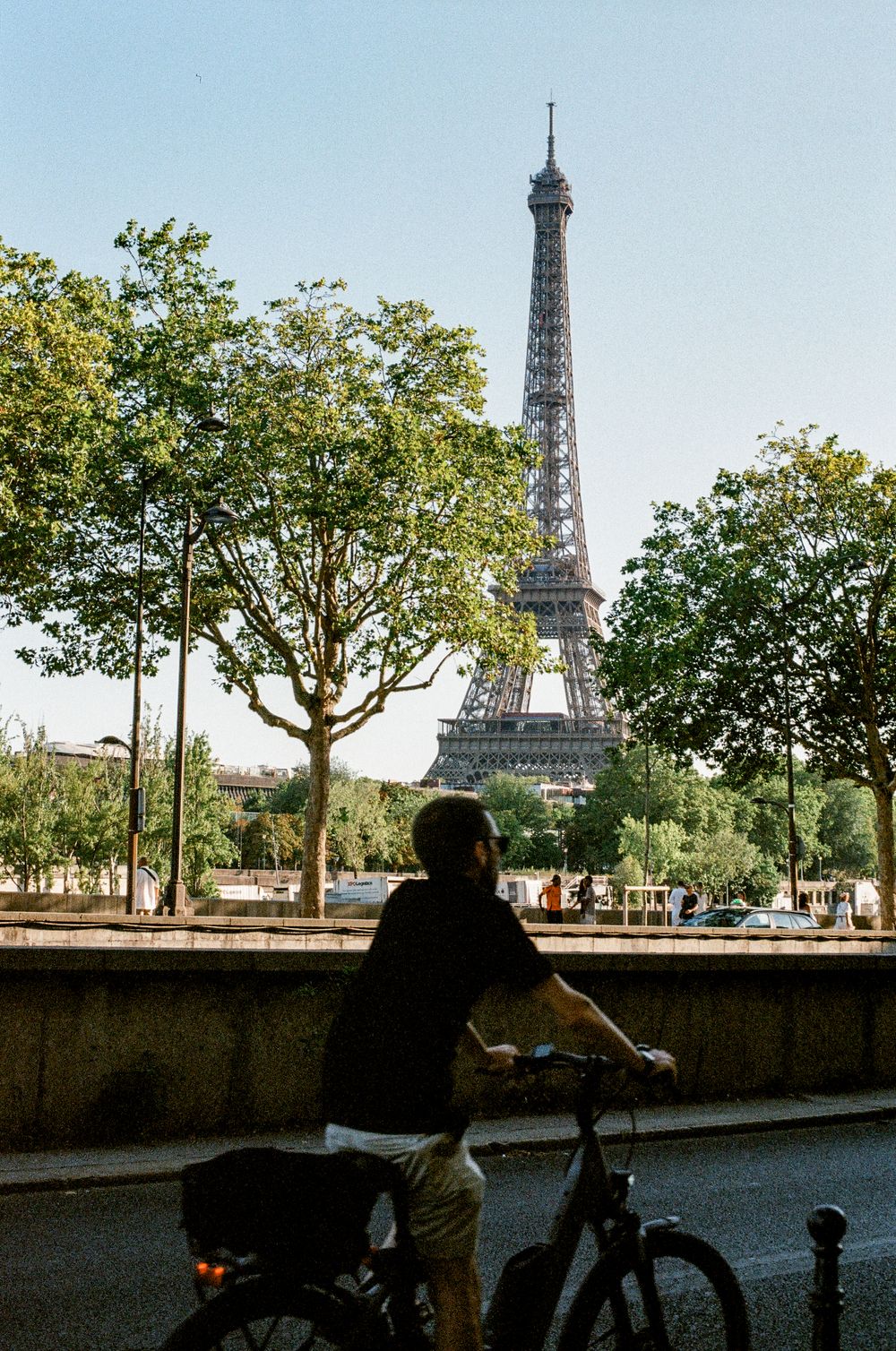 Cyclist rides along a Parisian street with the iconic Eiffel Tower rising above lush green trees on a clear sunny day.