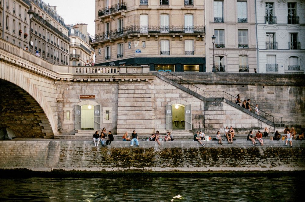 People relaxing on the banks of the Seine River in Paris, France, with a historic stone bridge and Haussmann buildings.