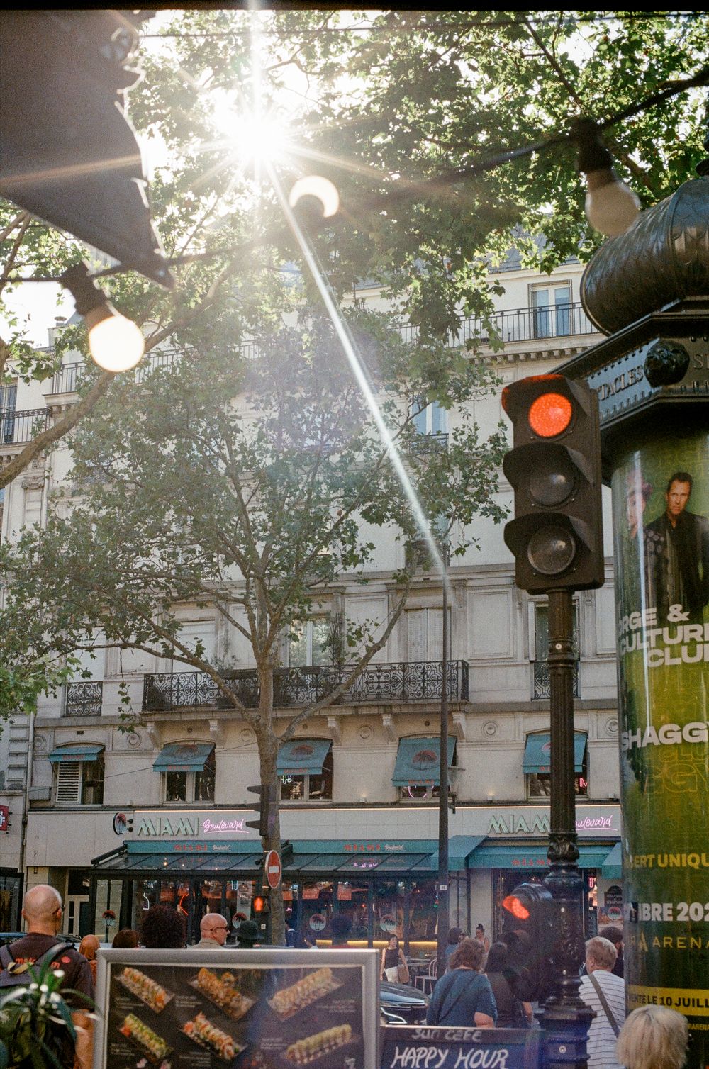 Sunlit Paris street scene with red traffic light, trees, Haussmann buildings, and Happy Hour sign on a busy boulevard.