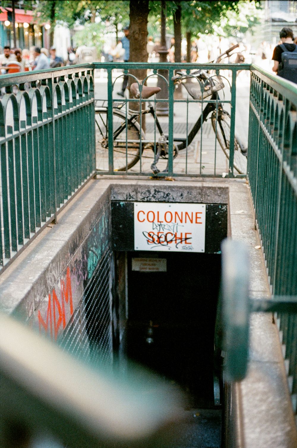 Underground entrance in Paris with green railings, a bicycle, graffiti, and a sign reading Colonne Seche.