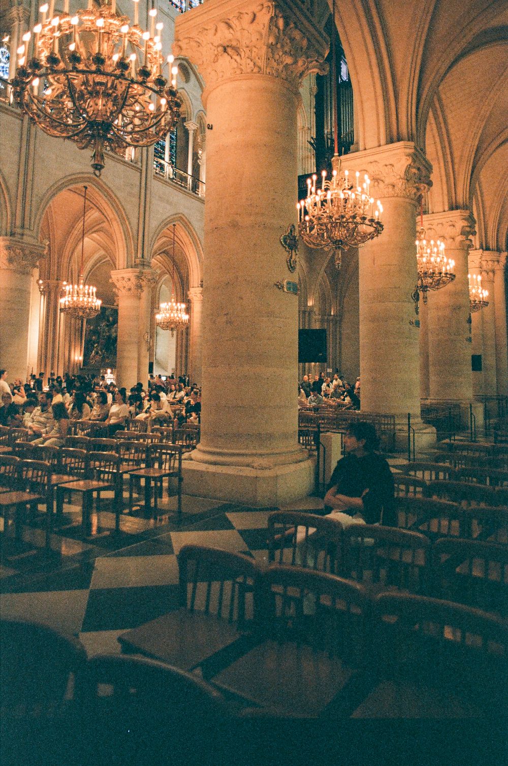 Interior of Notre-Dame Cathedral featuring grand chandeliers, Gothic arches, stone columns, and visitors seated in wooden chairs.