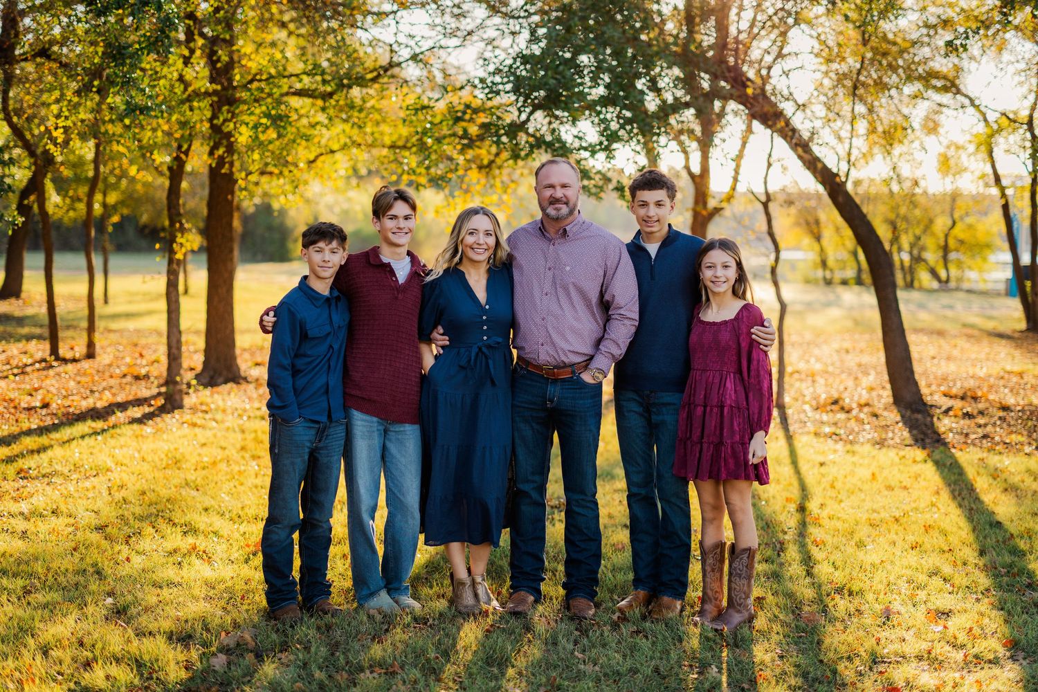 Family of six posing outdoors in golden autumn light among green trees on a sunny day.
