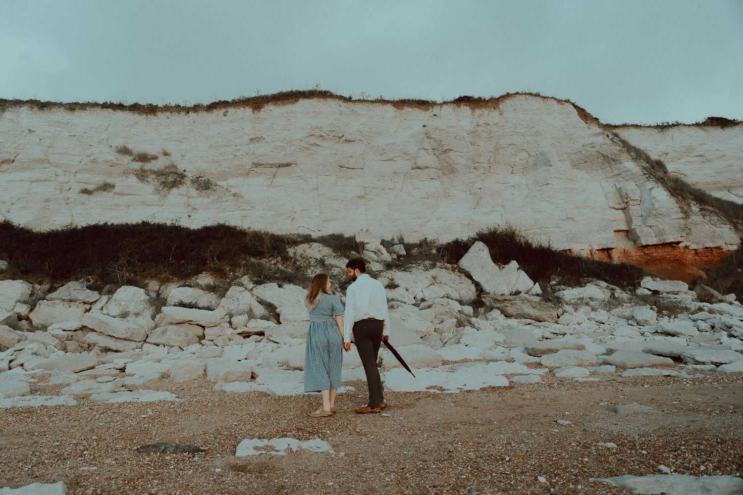 Couple standing together on a rocky beach beneath towering white chalk cliffs on an overcast day.