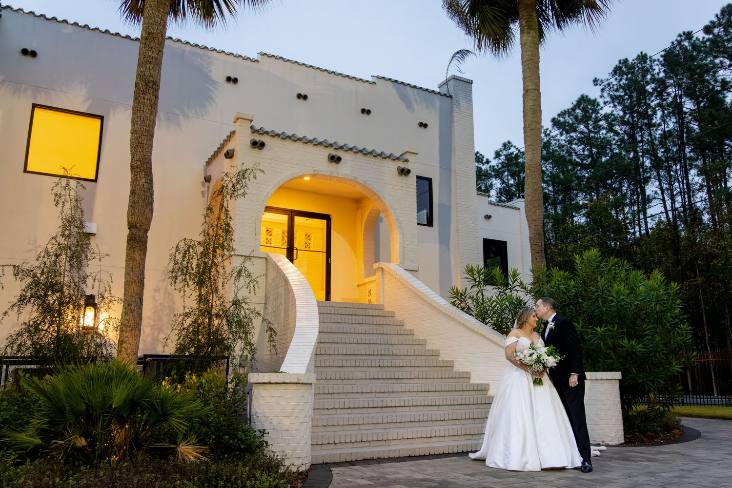 Bride and groom share a romantic kiss on elegant white staircase outside a stunning Spanish-style wedding venue at dusk.