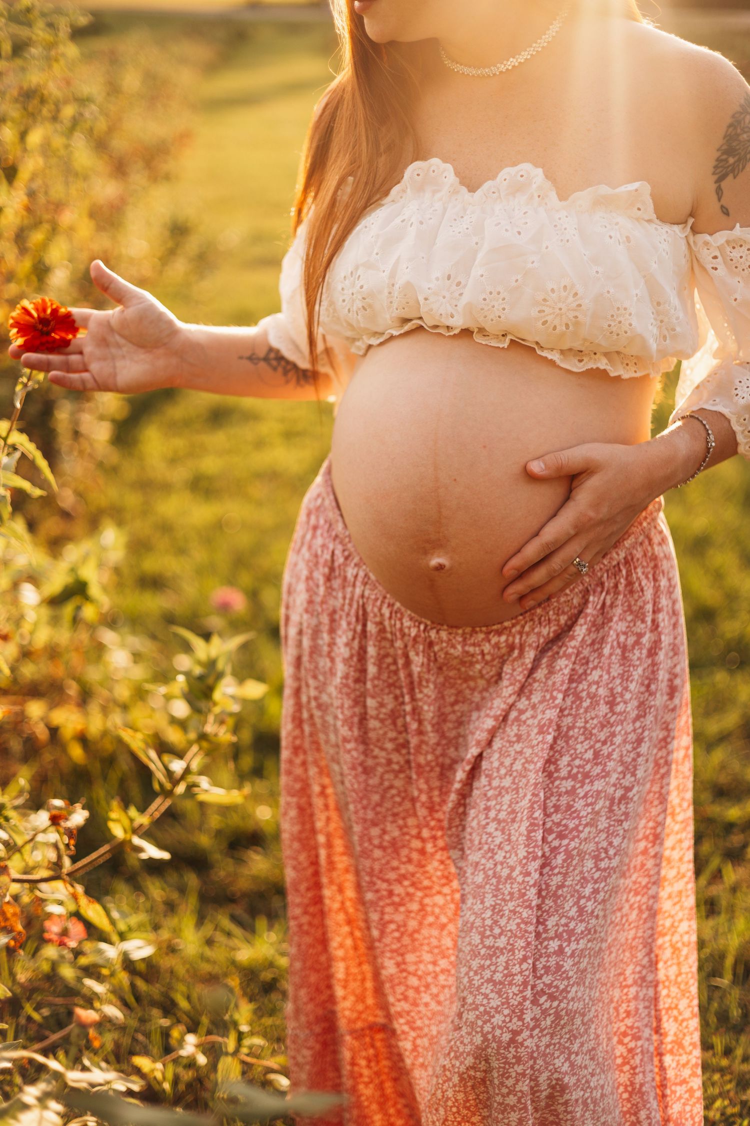 Pregnant mother holding a red flower and gently resting her hand on her baby bump during a golden-hour maternity photography session.