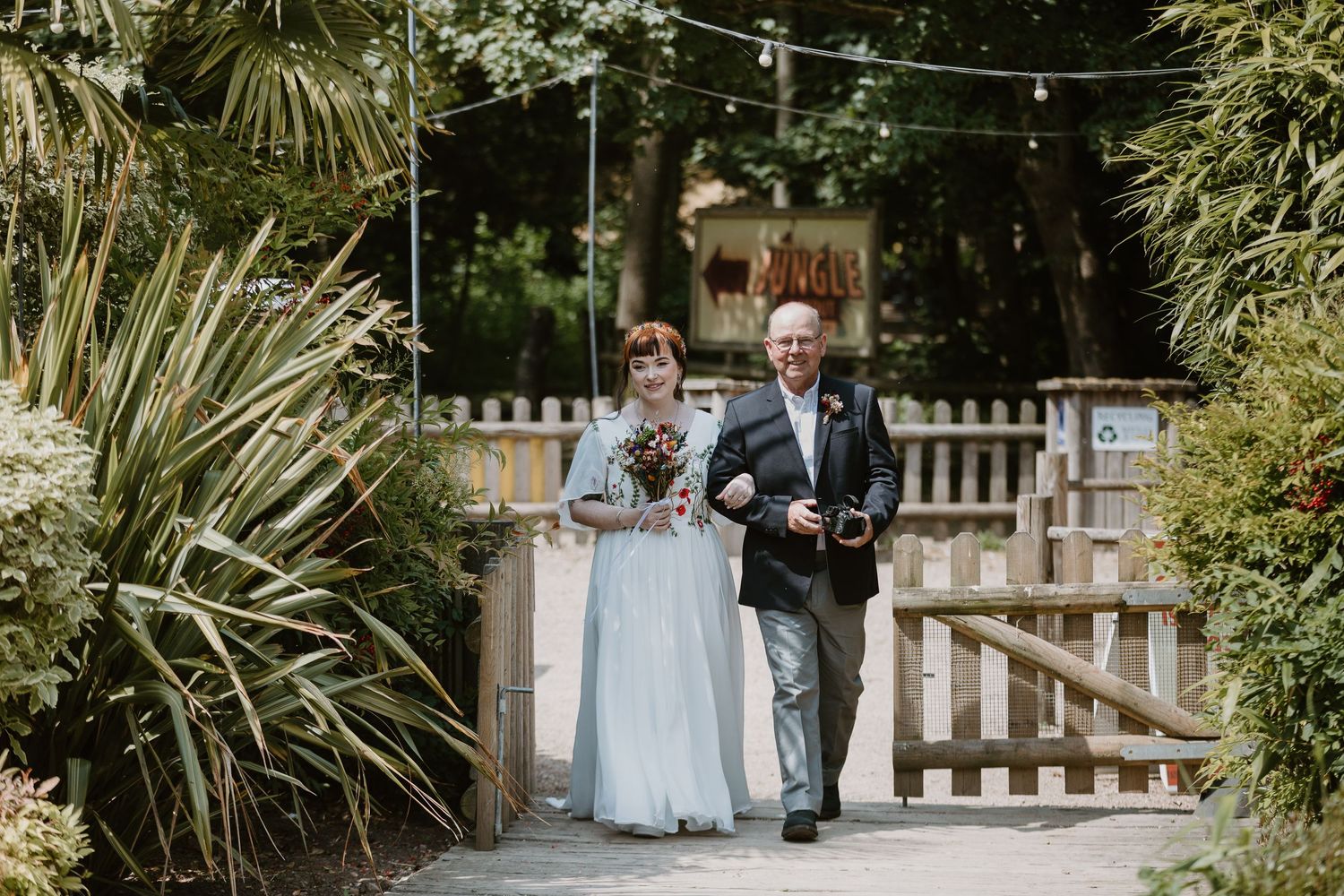 Bride in blue gown walks with escort through garden gate at outdoor wedding venue.