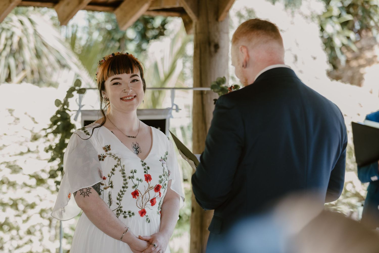 Bride smiling at groom during outdoor wedding ceremony, wearing white floral embroidered dress under wooden arbor.