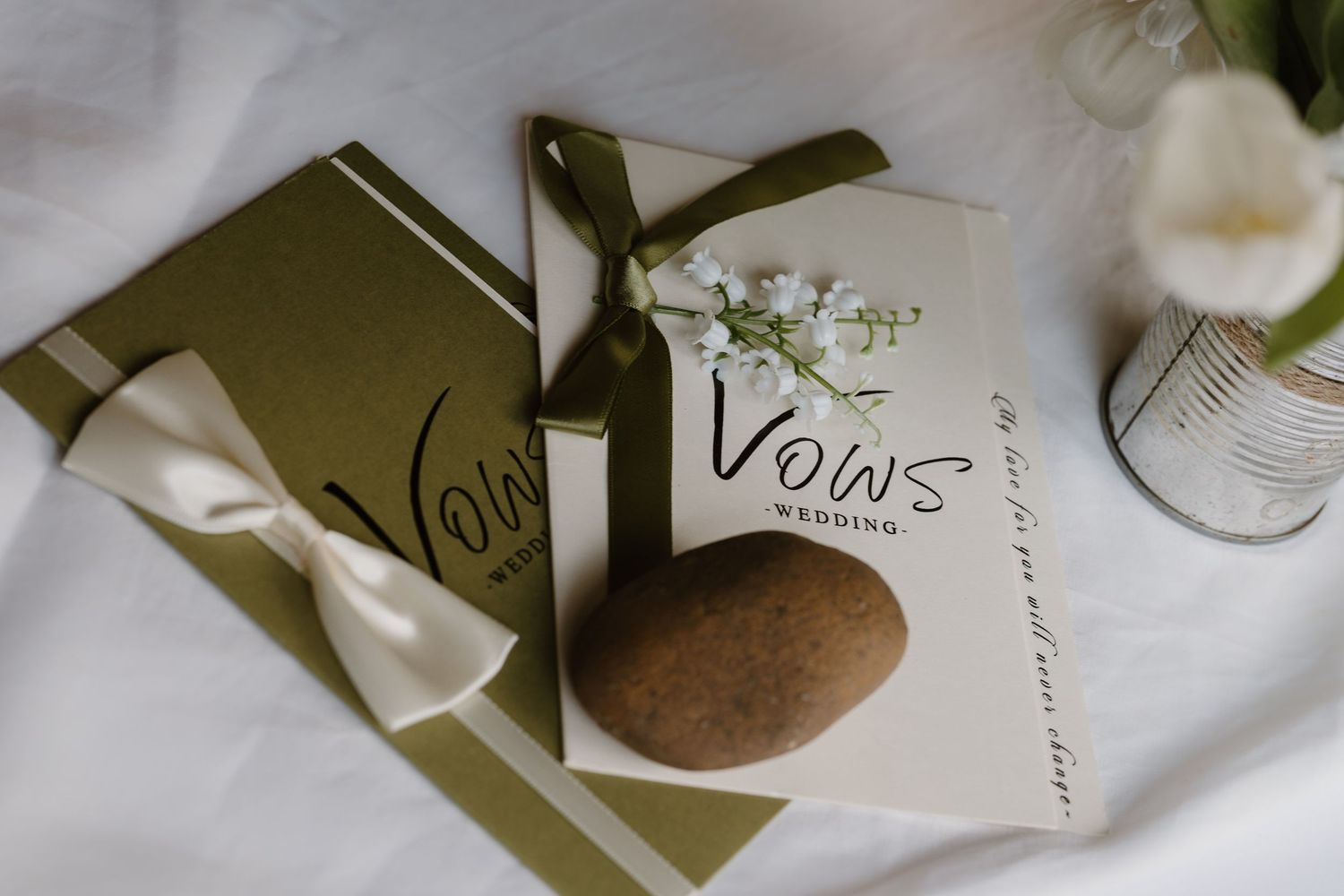Elegant olive green wedding vow books with white ribbon bow and floral accents on white background.
